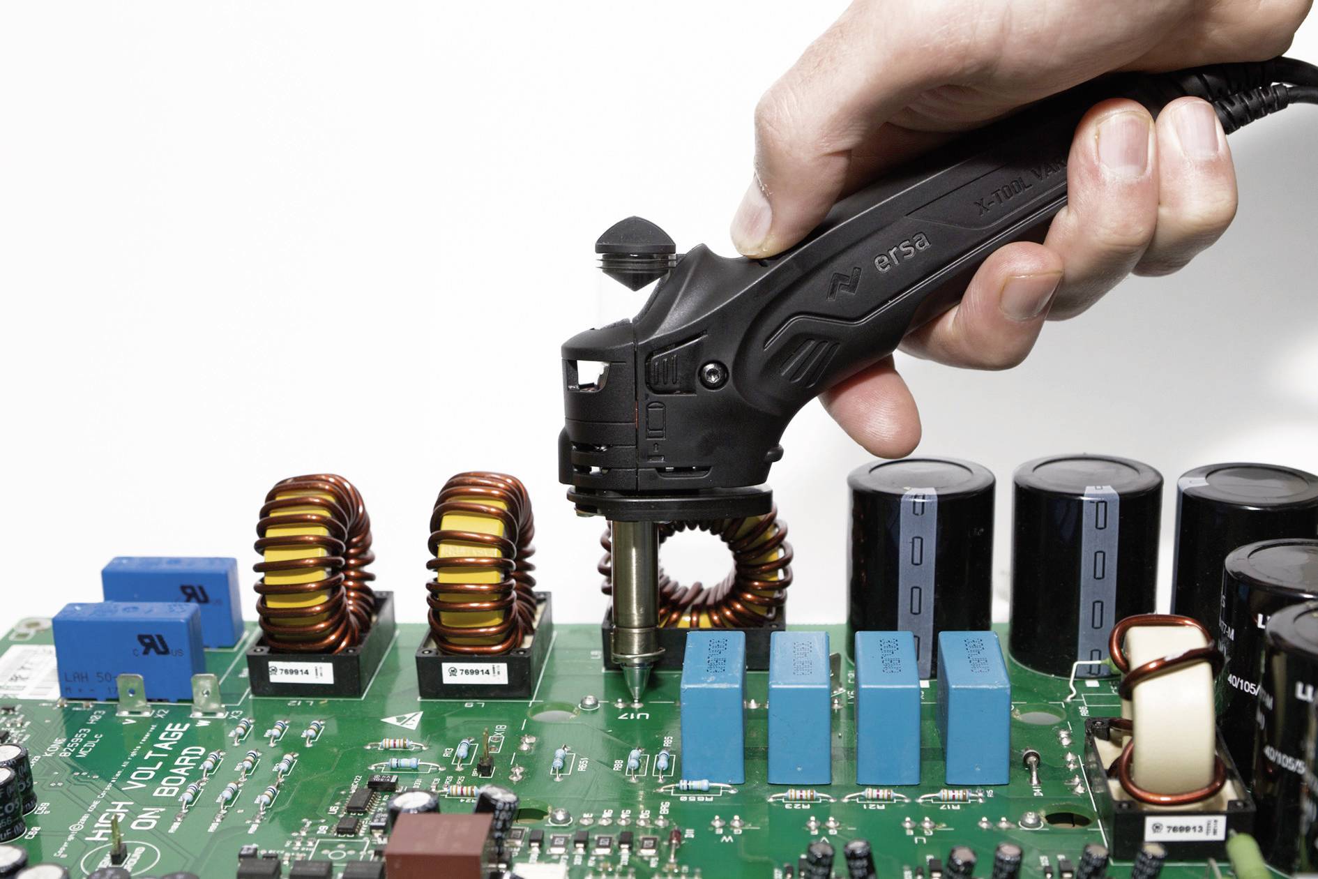 A hand holds a soldering iron and works on a green circuit board, with electronic components such as coils and capacitors visible.