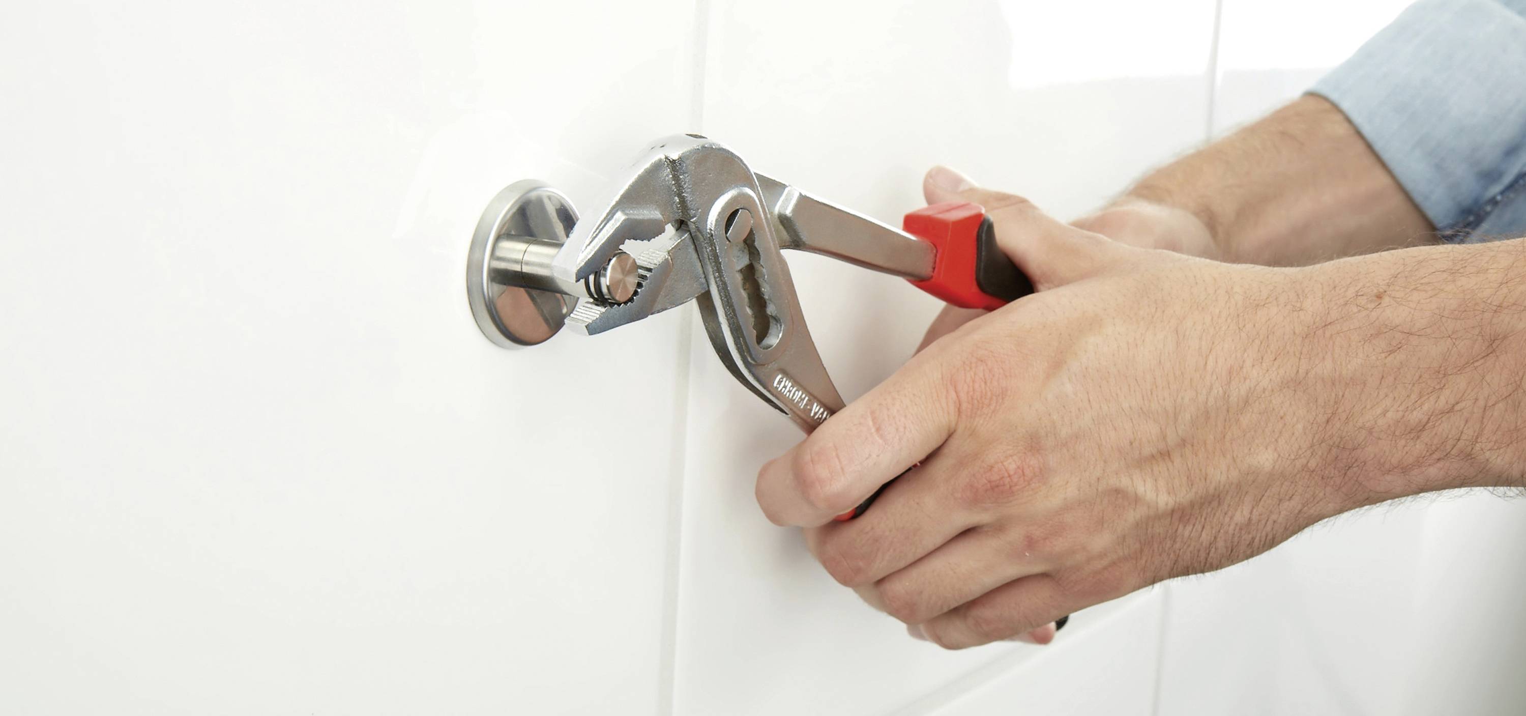 A person is repairing a fitting with a pipe wrench, which is placed on a pipe section against a white wall.