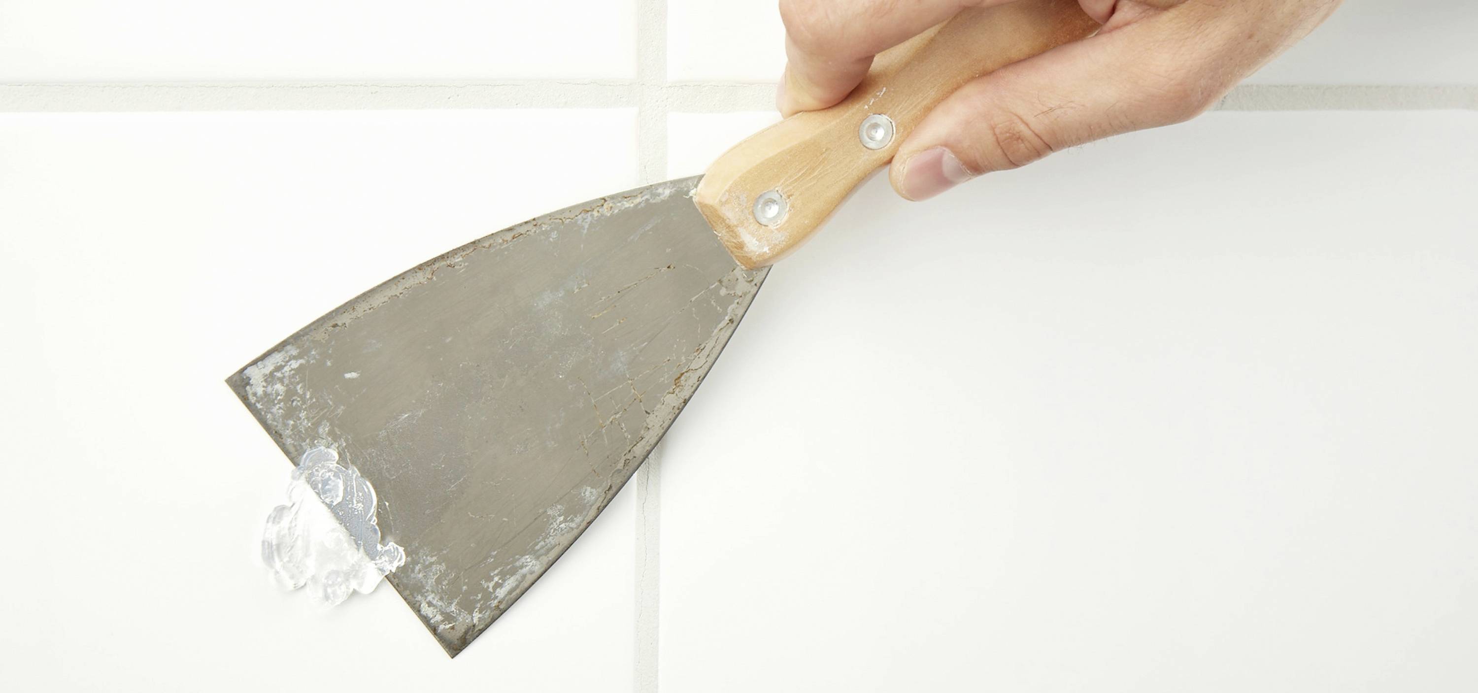 A hand is holding a scraper and removing silicone from a tile.