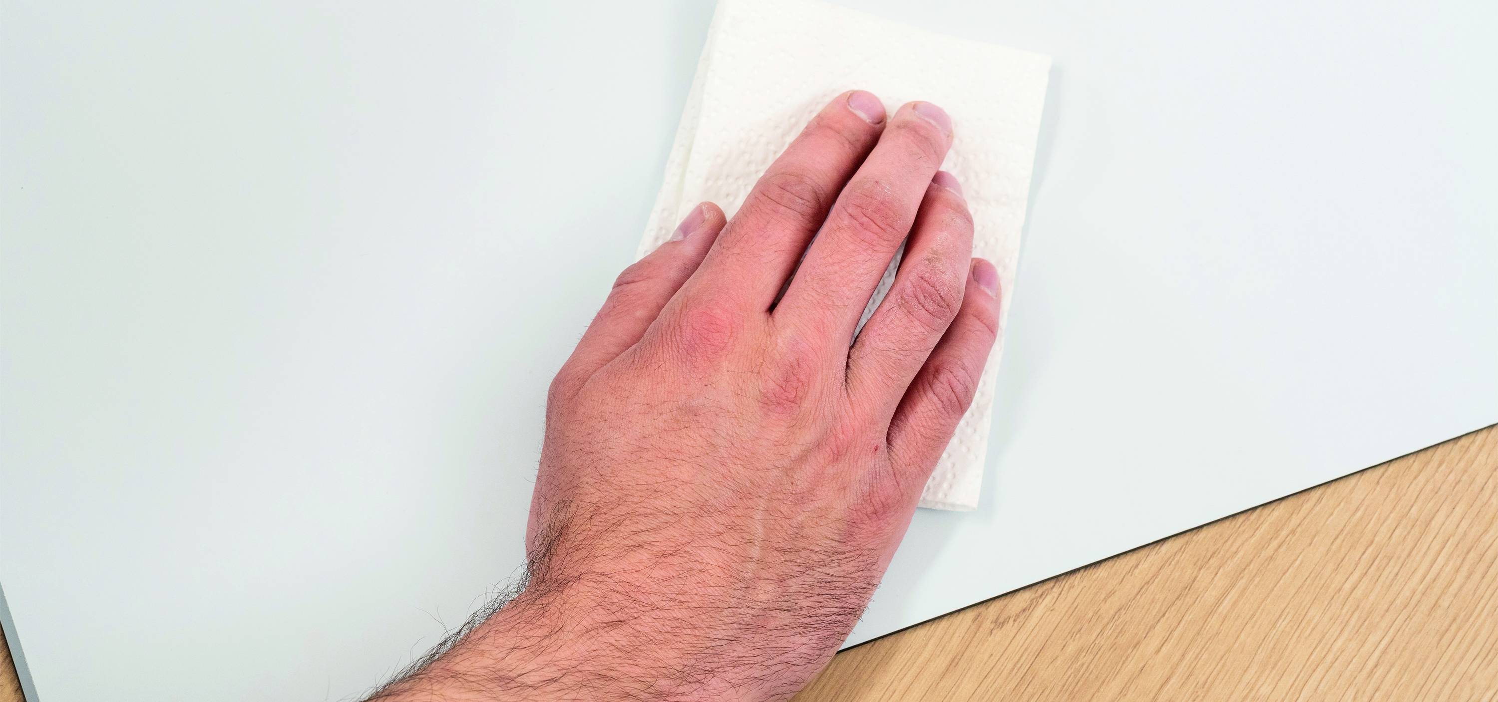 A hand wipes a smooth, light surface with a white cloth. Part of a wooden surface is visible underneath.