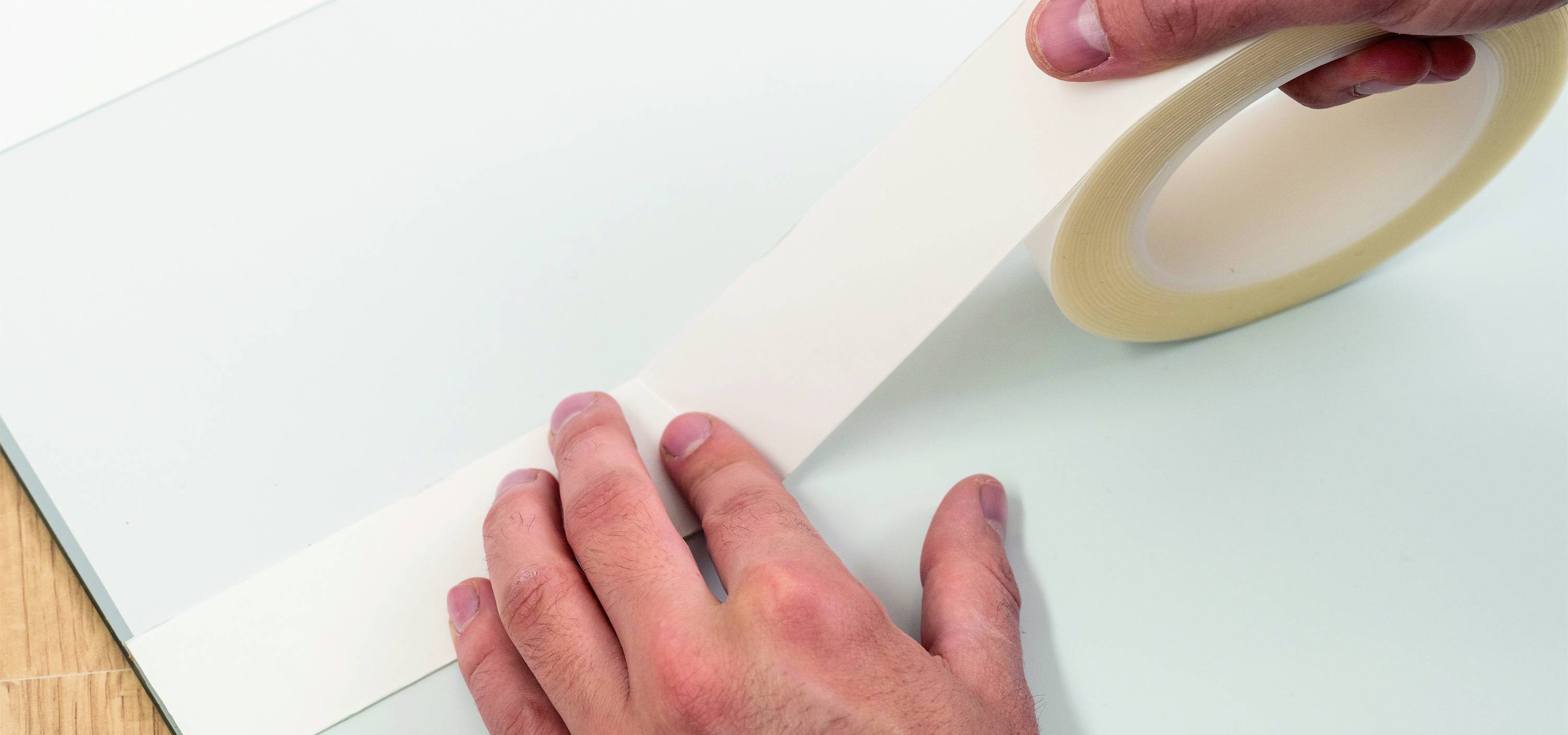 Two hands are sticking a piece of adhesive tape onto a smooth, light-coloured surface.