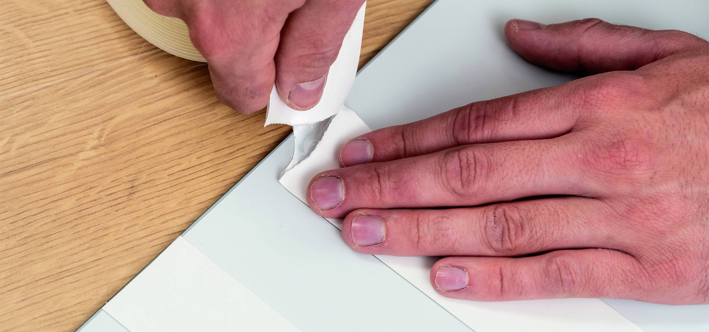 A person is applying masking tape to the edge of a surface on a table. The hands demonstrate careful workmanship as the tape is being unrolled.