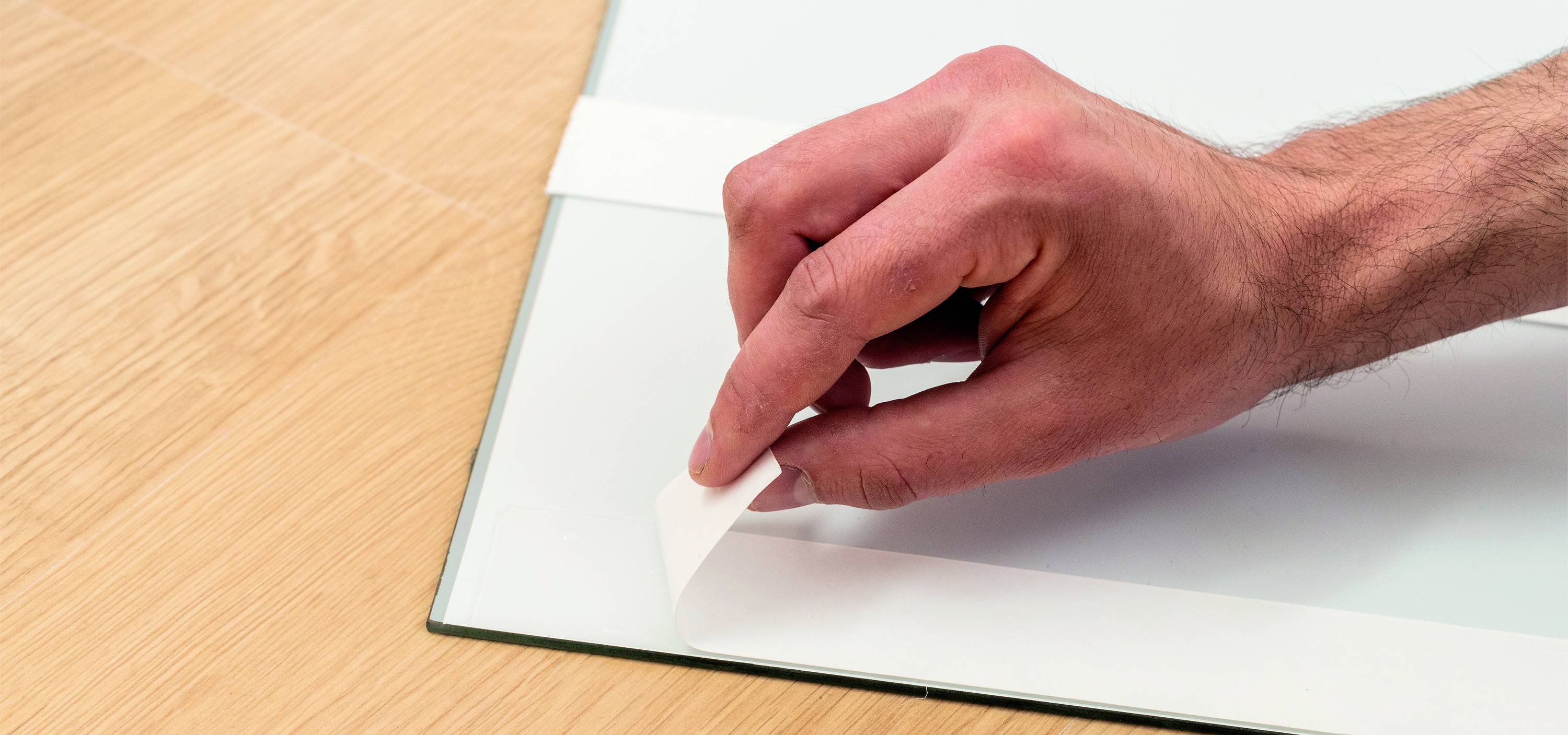 A hand peels a strip of adhesive tape from a glass surface, lying on a wooden floor.