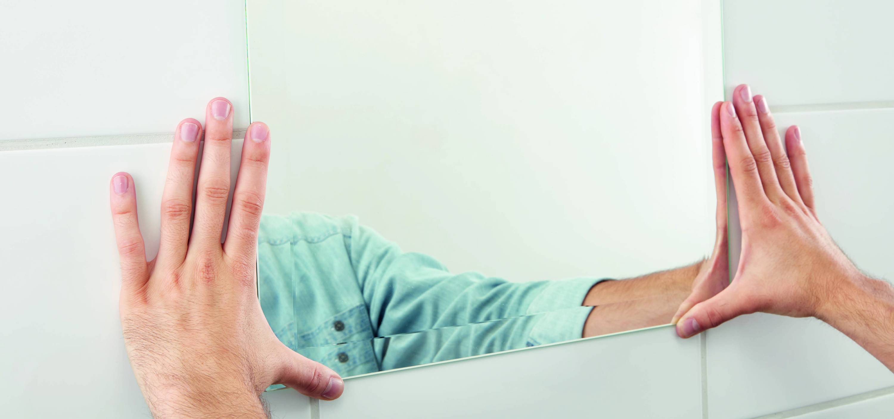 A person is aligning a mirror on a tiled wall. Only hands and part of the upper body are visible.