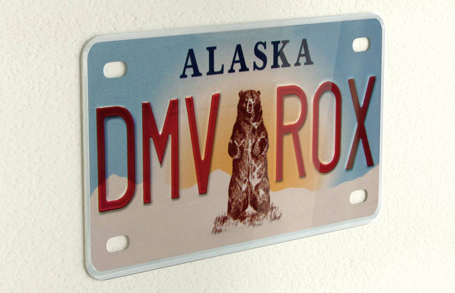 Vehicle registration plate from Alaska with the letters 'DMV ROX' and a bear image in the centre, set against a mountainous backdrop.