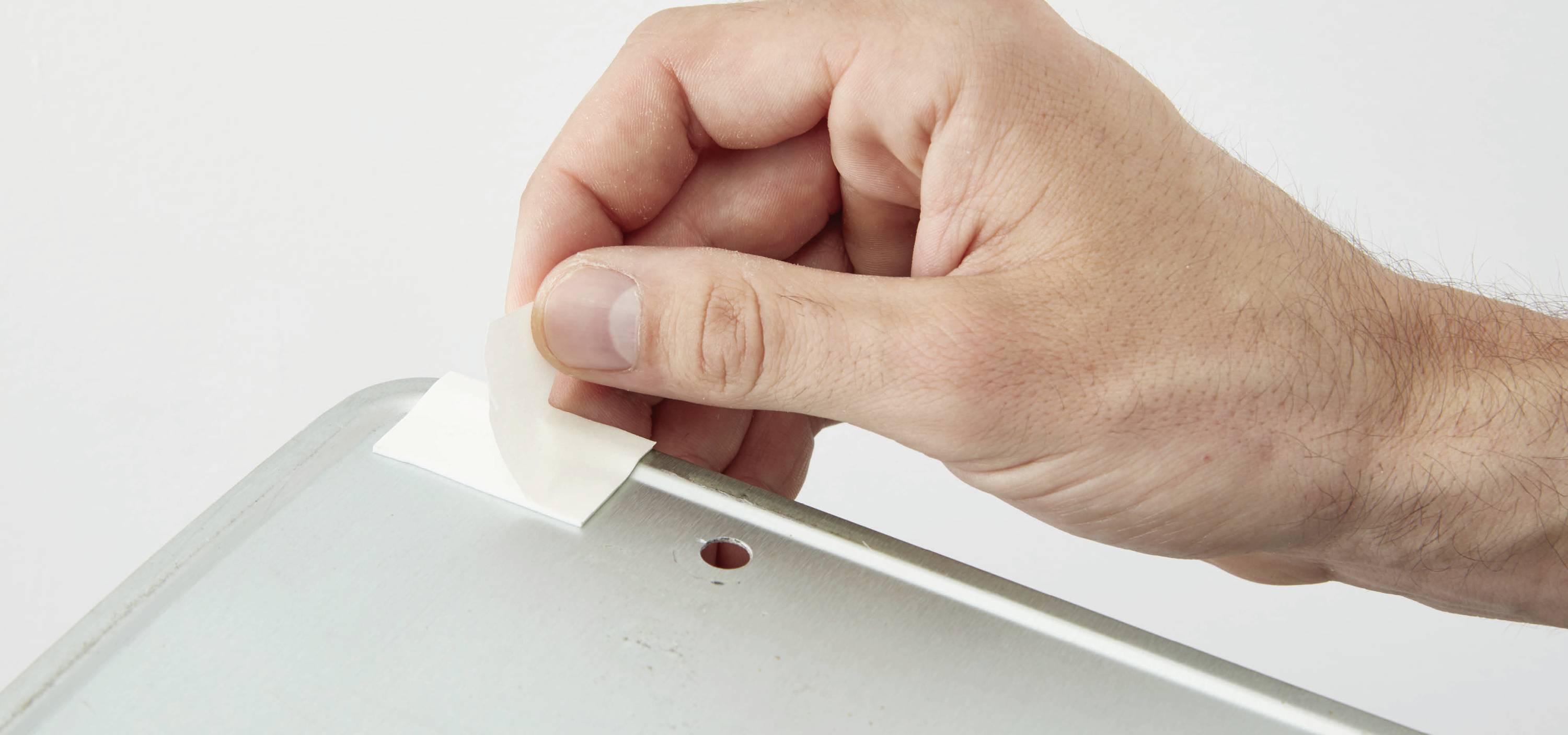 A hand peels a strip of adhesive tape from a metallic surface.