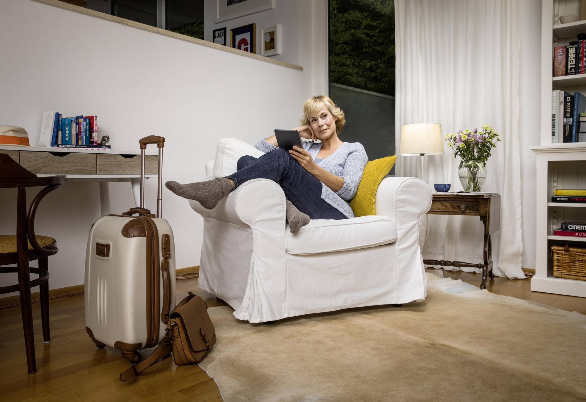 A woman sits relaxed in an armchair, reading on a tablet. A suitcase stands beside her. The room is cosily furnished.