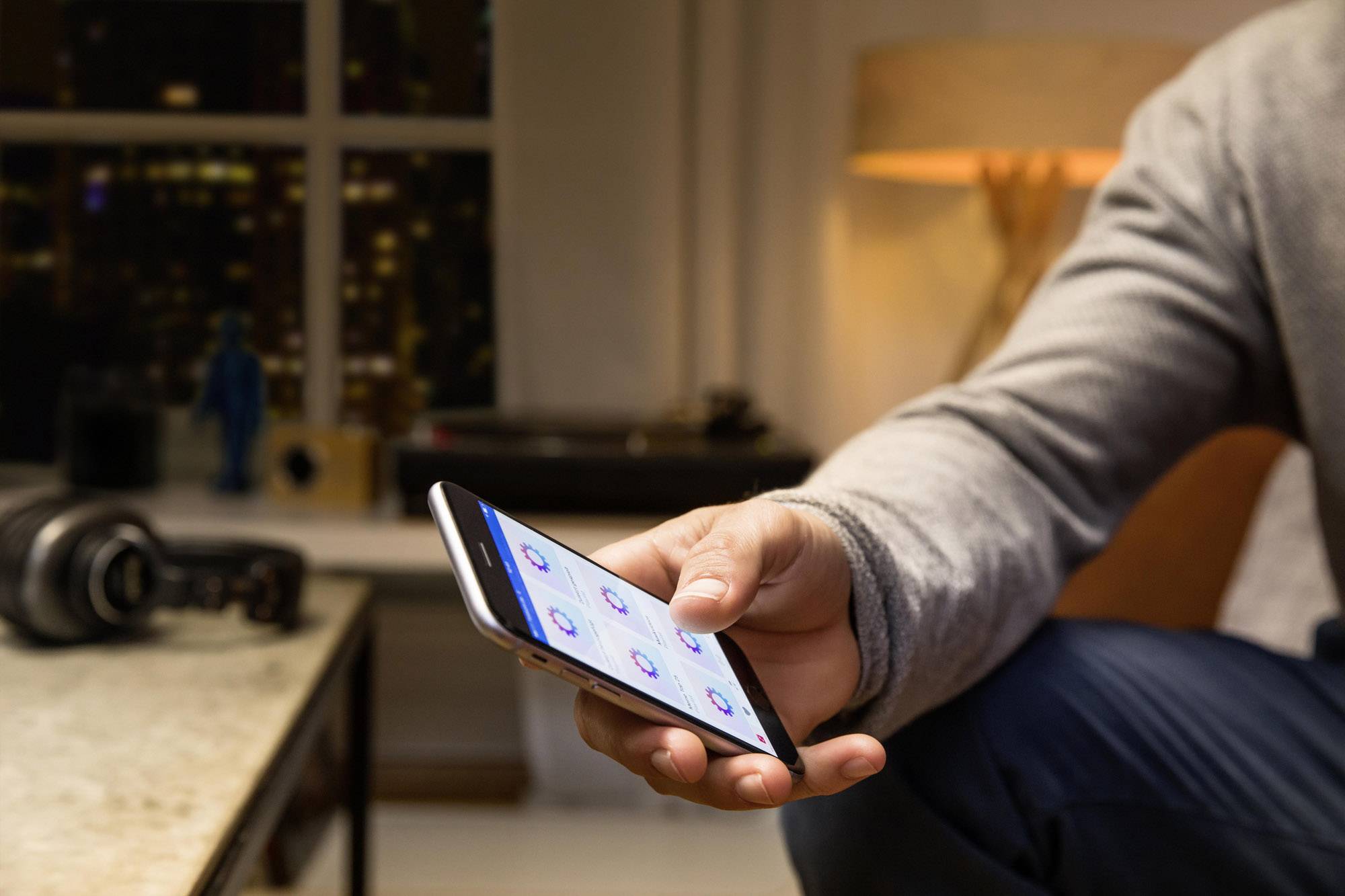 A person is holding a smartphone, the screen displaying multiple app icons. A cosy living room scene is visible in the background.