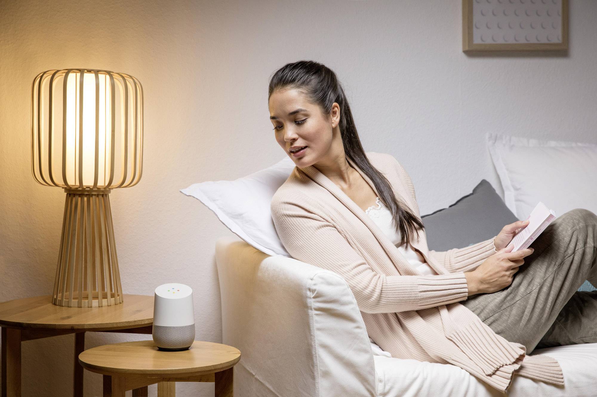 A woman is sitting relaxed on a sofa, reading a book and interacting with a voice assistant on a table beside her.