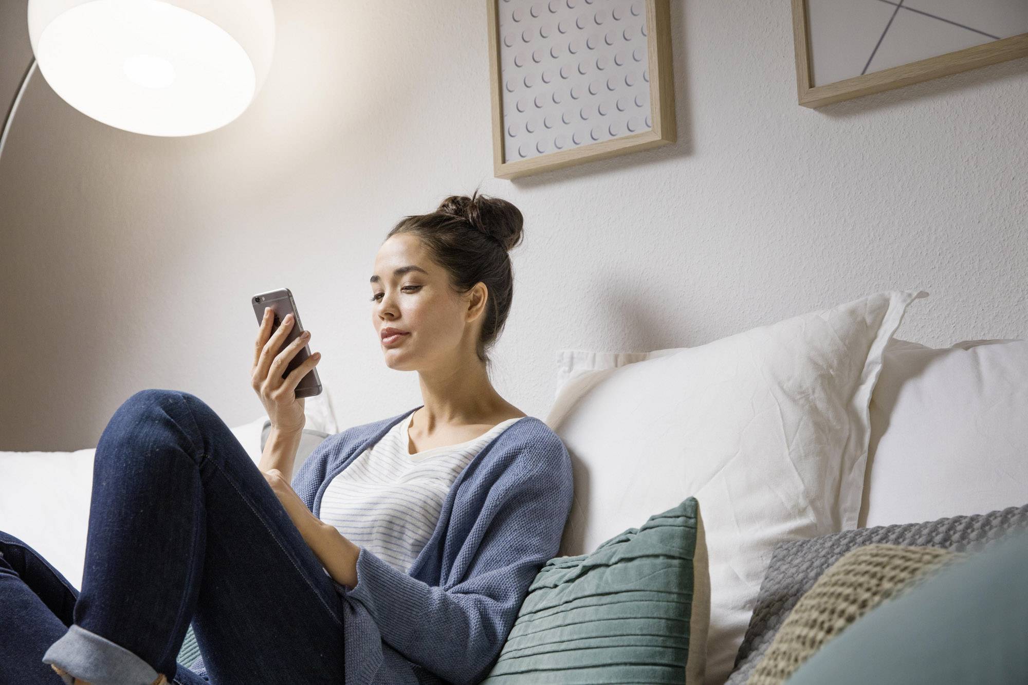 A woman sits relaxed on a sofa, looking at her smartphone. She is wearing casual clothing, with picture frames in the background.