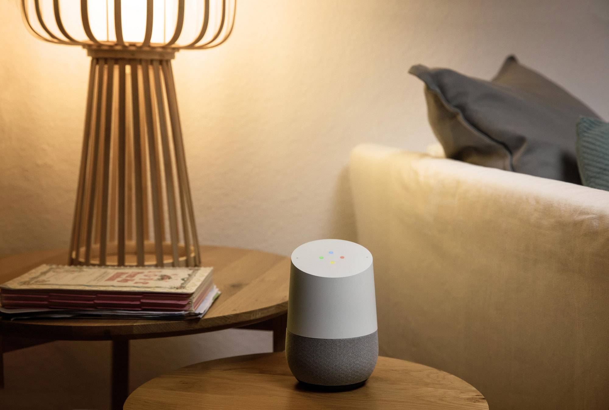 A smart speaker sits on a wooden table next to a glowing lamp and a sofa. Cushions are visible in the background.