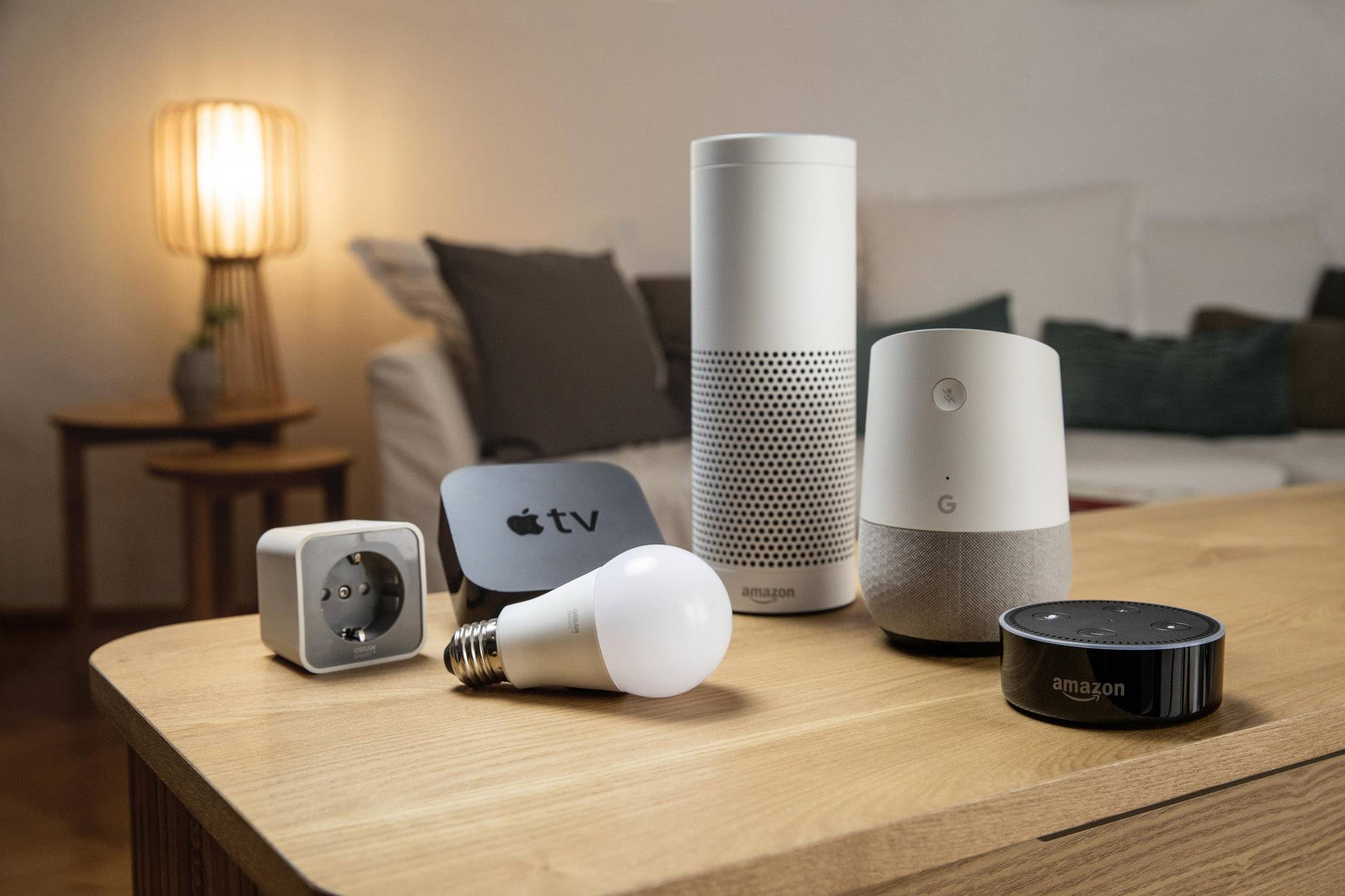 Several smart home devices on a table, including a speaker, an LED light bulb, and a streaming device, in front of a sofa.