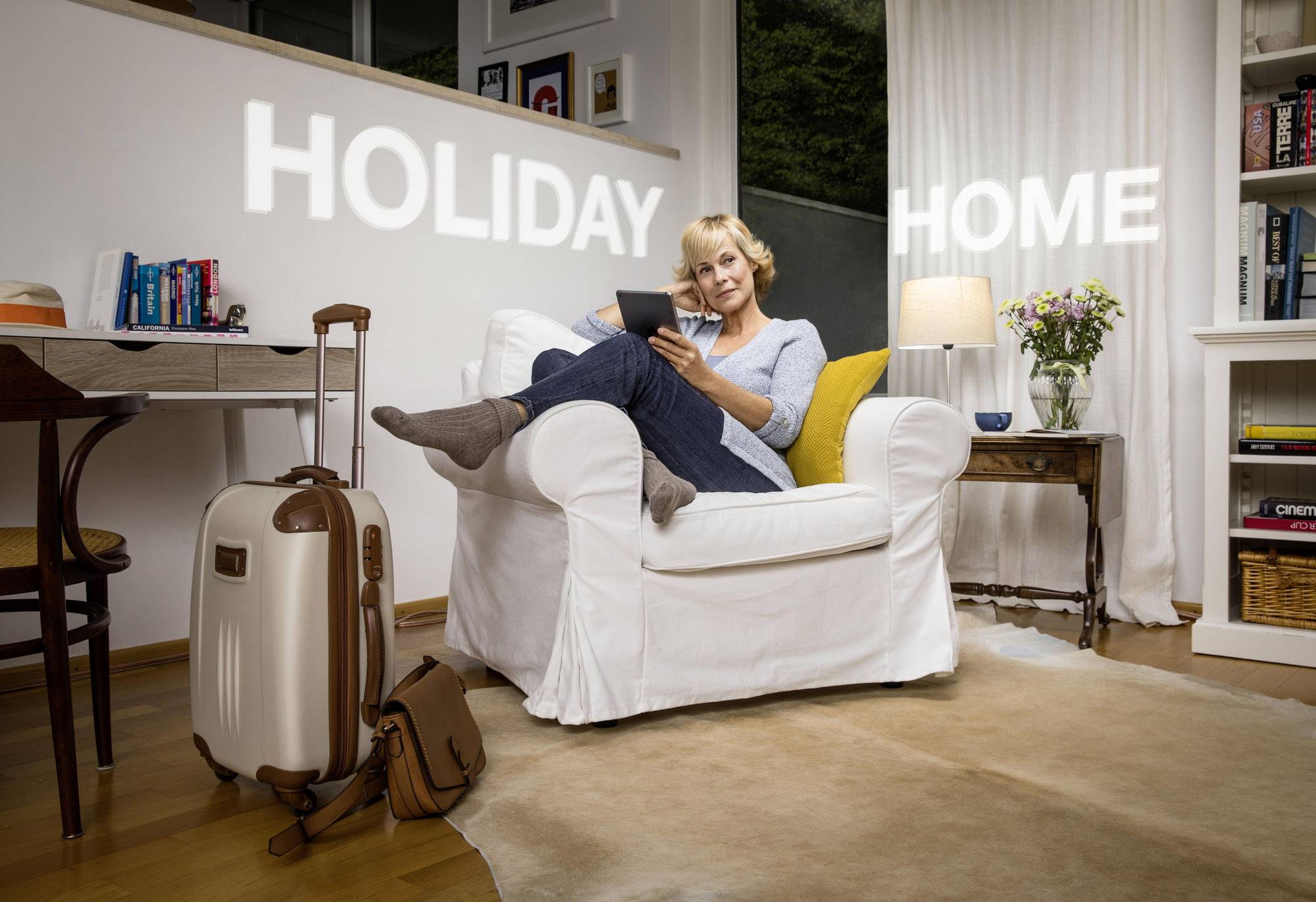 A woman sits relaxed on a white armchair with a tablet in her hand; 'Holiday Home' illuminates in the background.
