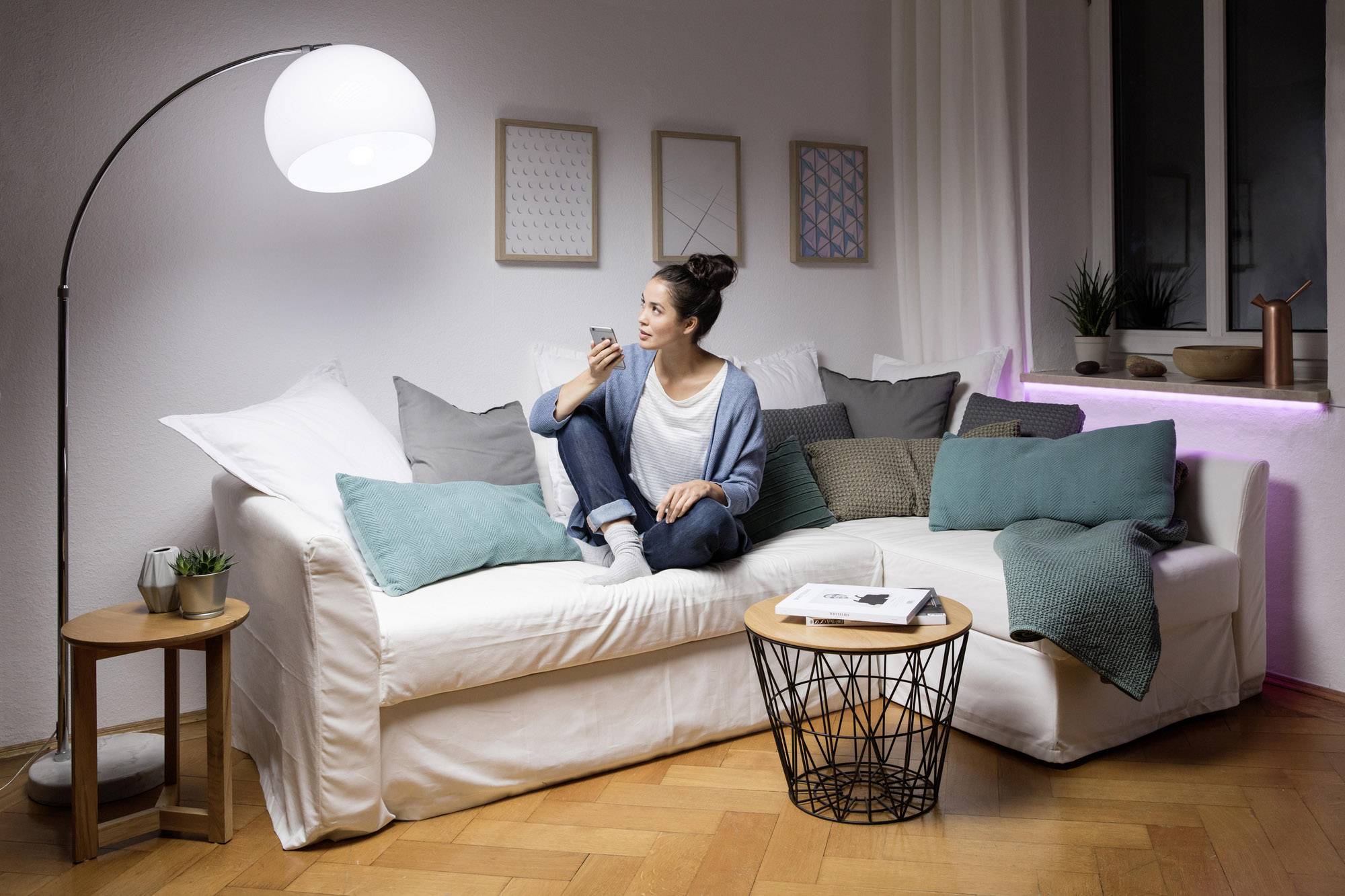 A person is sitting relaxed on a white sofa, surrounded by cushions. They are holding a smartphone. The room is decorated in a modern and cosy style.