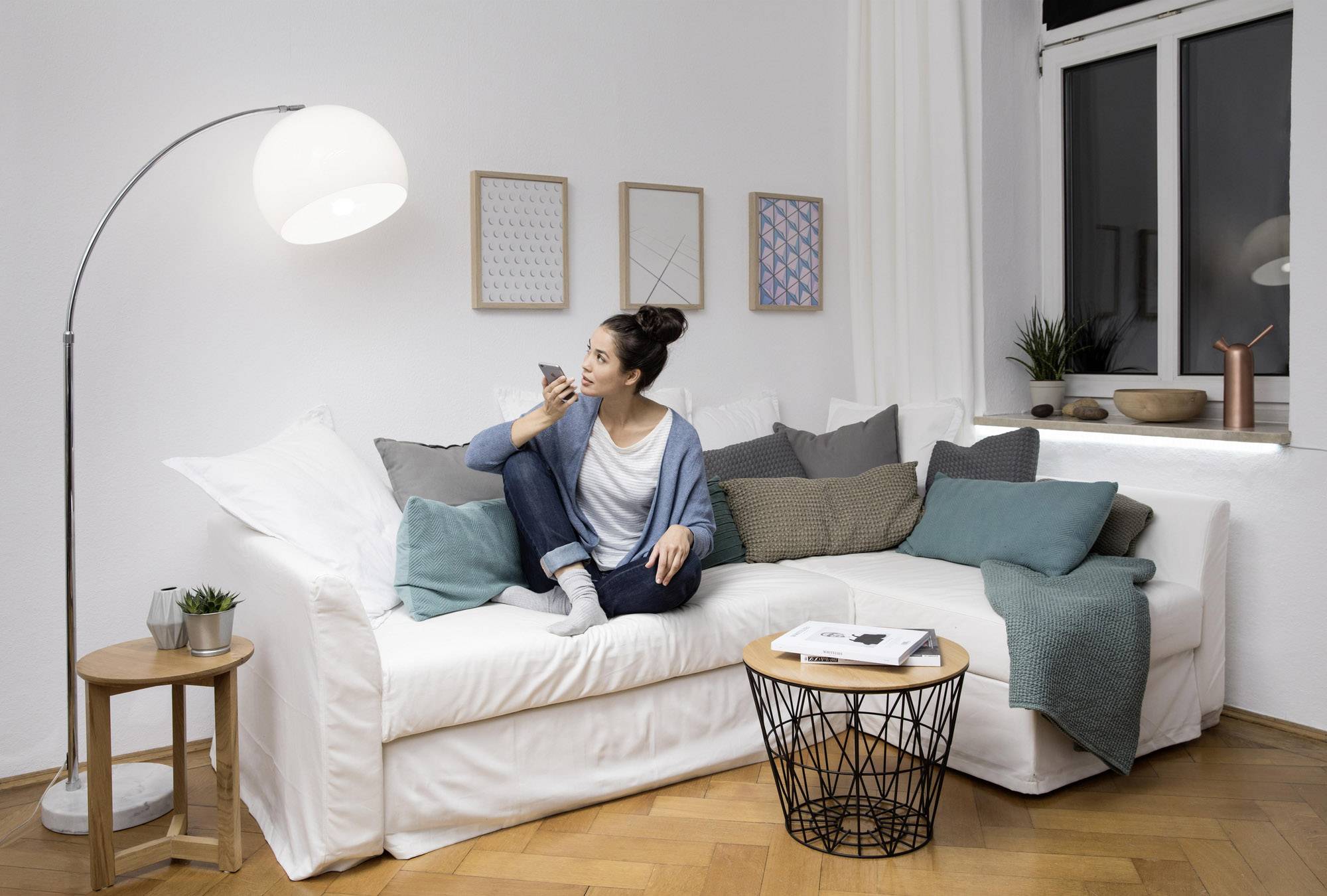 A woman sits relaxed on a white sofa, holding a mobile phone and gazing thoughtfully to the side. Modern furnished living room.