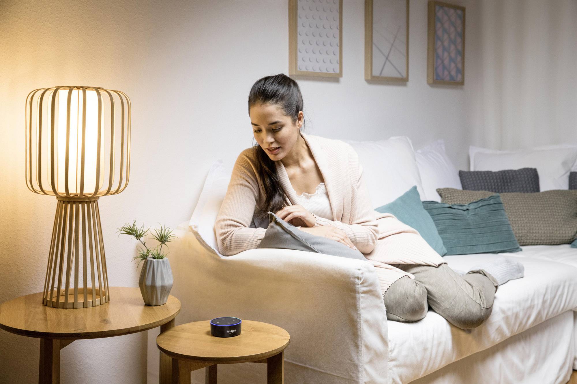 A woman is sitting on a sofa and speaking to a voice assistant device on a table. She is relaxed and the room is cosily lit.
