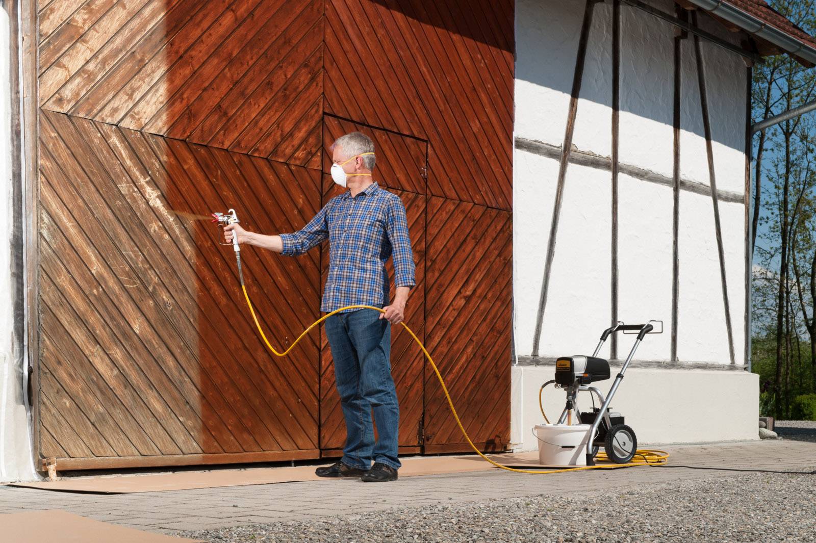A person is spraying paint onto a large wooden gate using a spray gun. The person is wearing a protective mask.