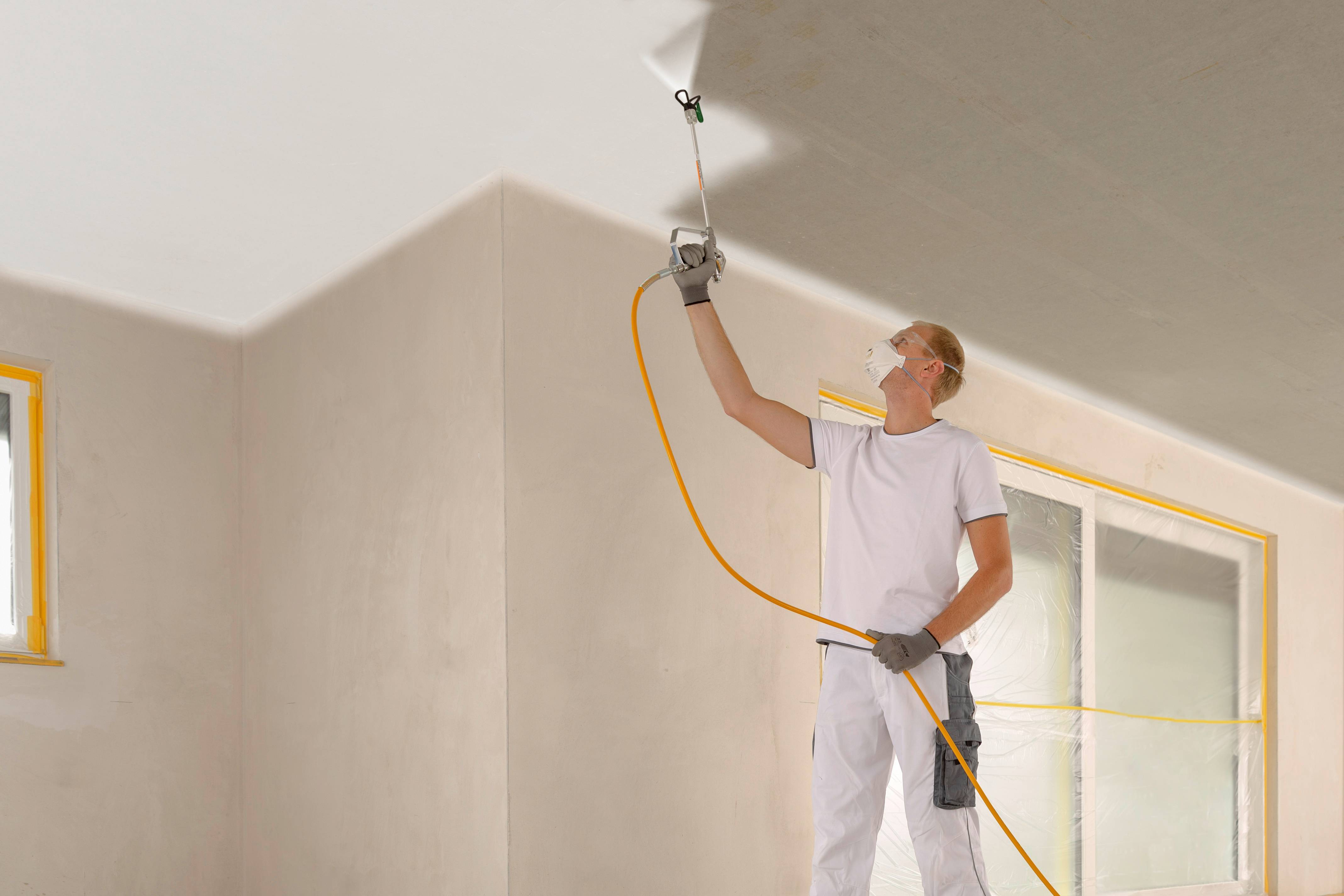 A painter in white workwear is spraying paint onto the ceiling with a long nozzle. Windows and walls are masked off with tape.