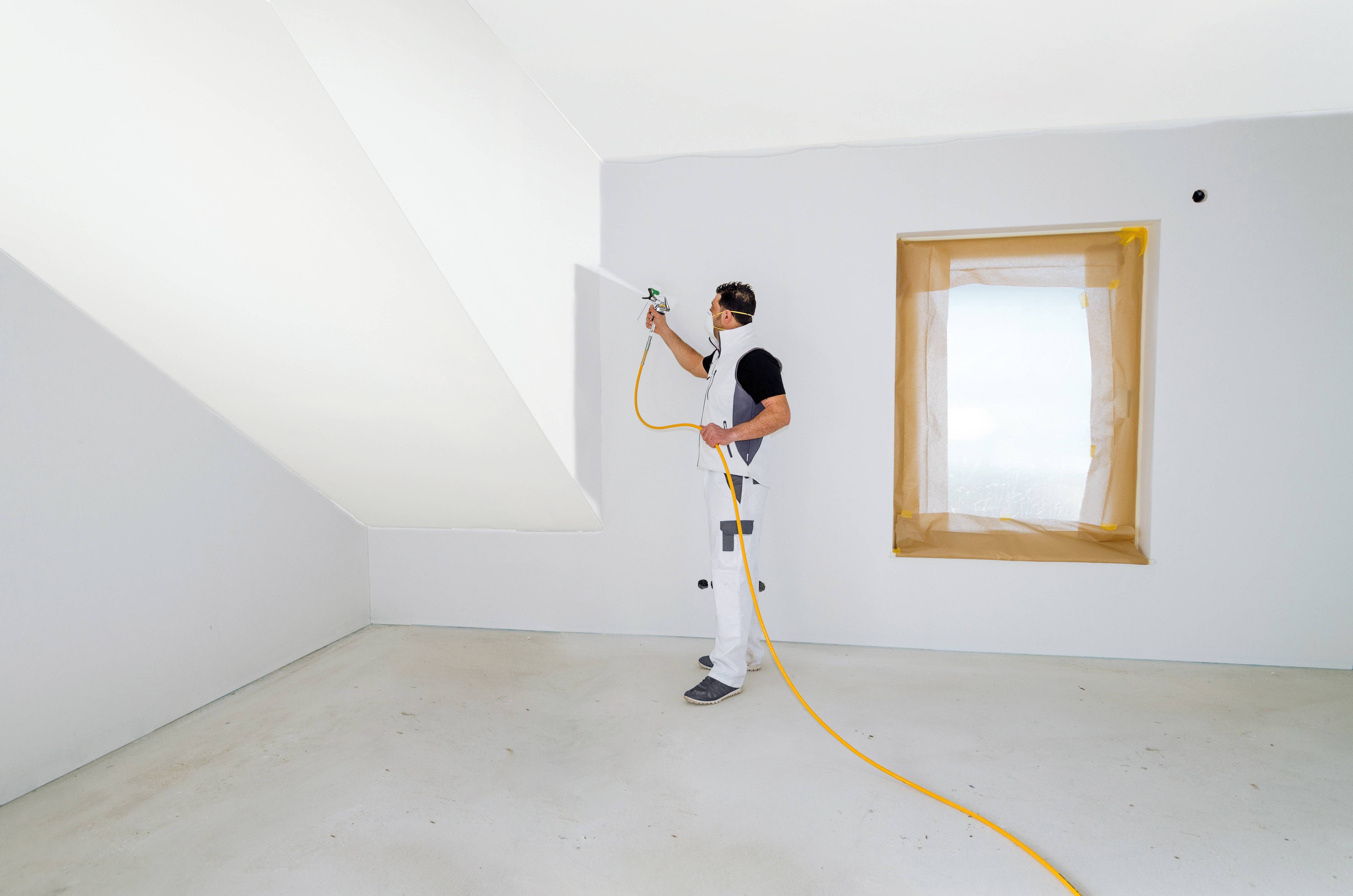 A painter in a white overall is spraying white paint onto a wall in an empty room. A window is covered.