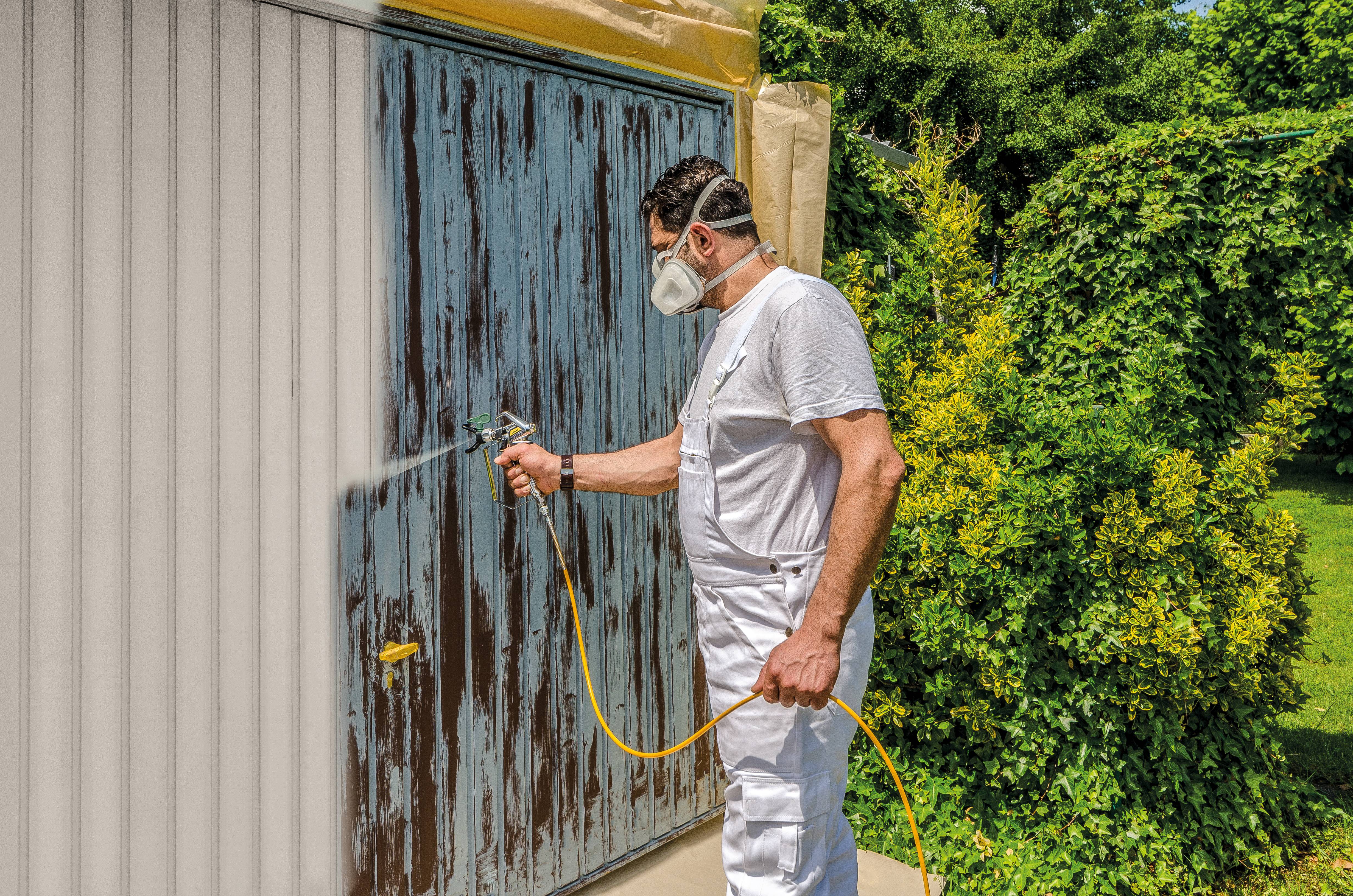 A man in white workwear is spray painting a garage door in a garden while wearing a respiratory mask.