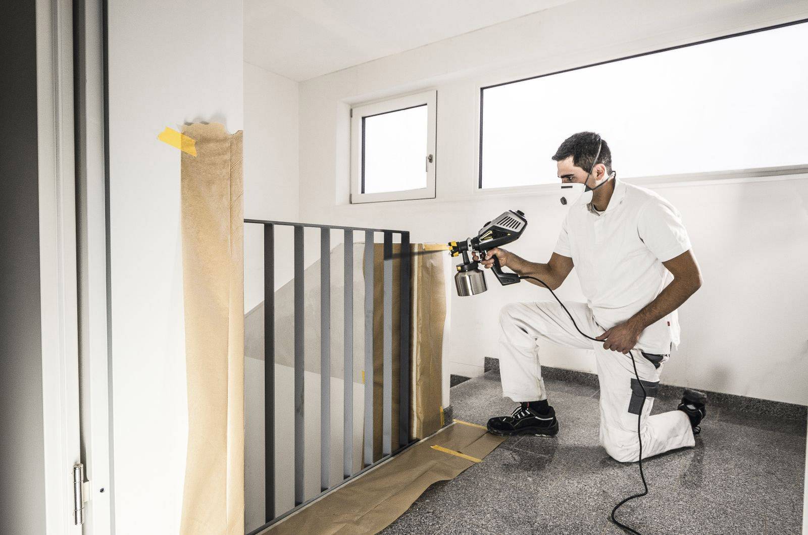 A man in protective clothing is spraying paint onto a stair banister. The room is prepared with dust sheets and coverings.