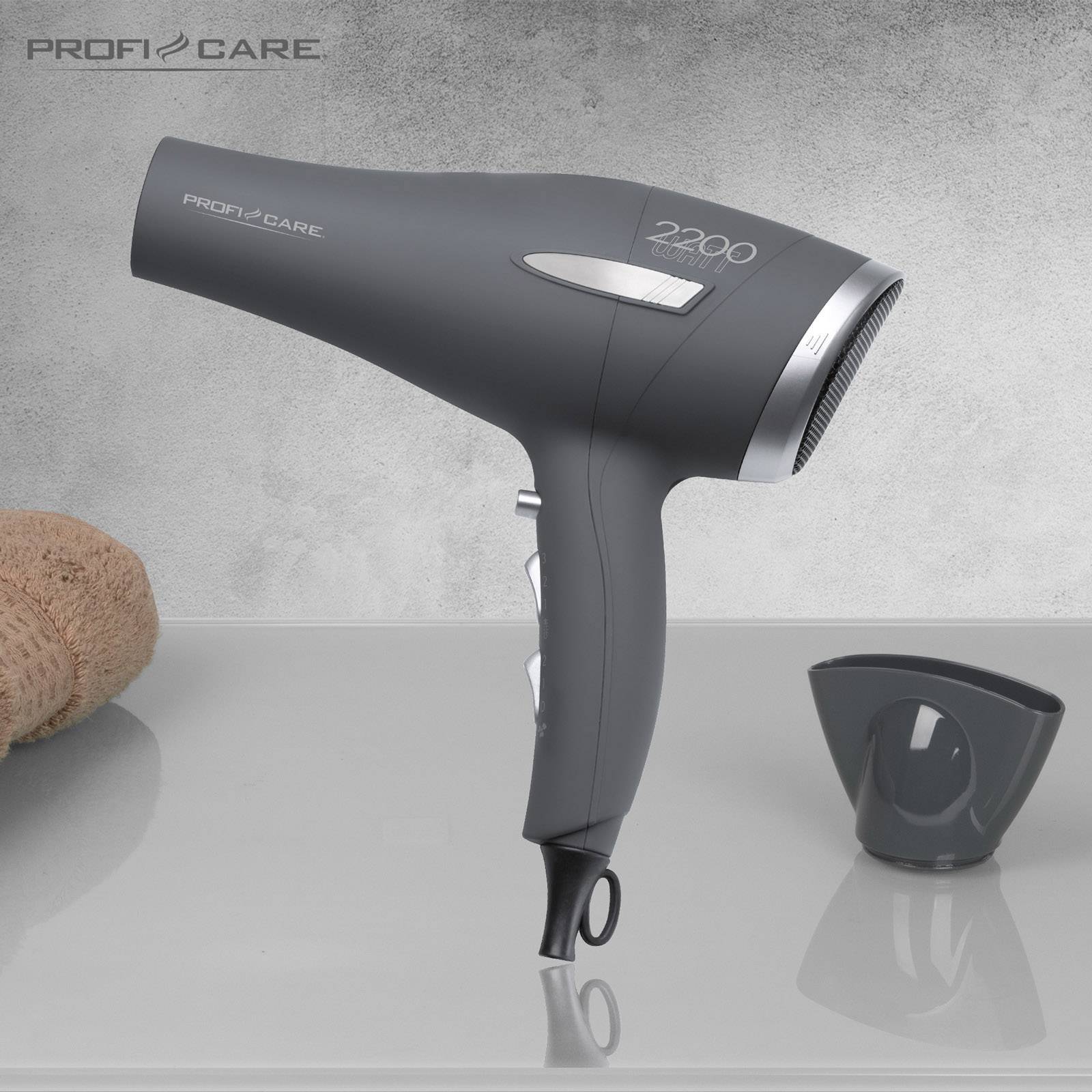 A grey hairdryer with a removable attachment lies on a grey surface, alongside a towel and a nozzle.