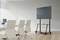 A conference room with a large screen on a mobile stand beside a table with chairs, suitable for presentations.