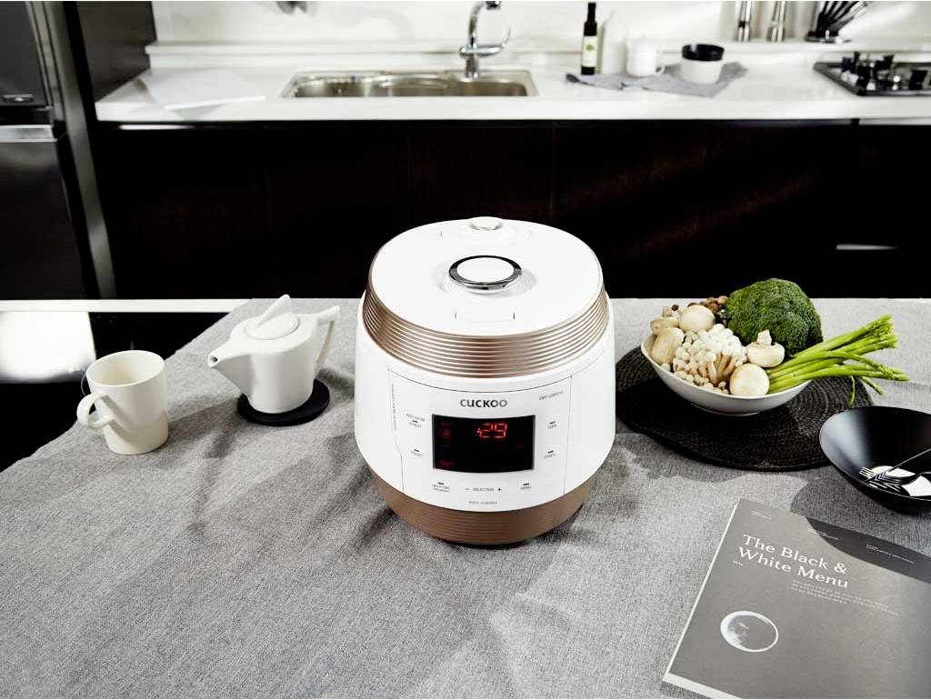 Electric pressure cooker on kitchen table next to teapot and bowl of vegetables; sink and kitchen worktop in the background.