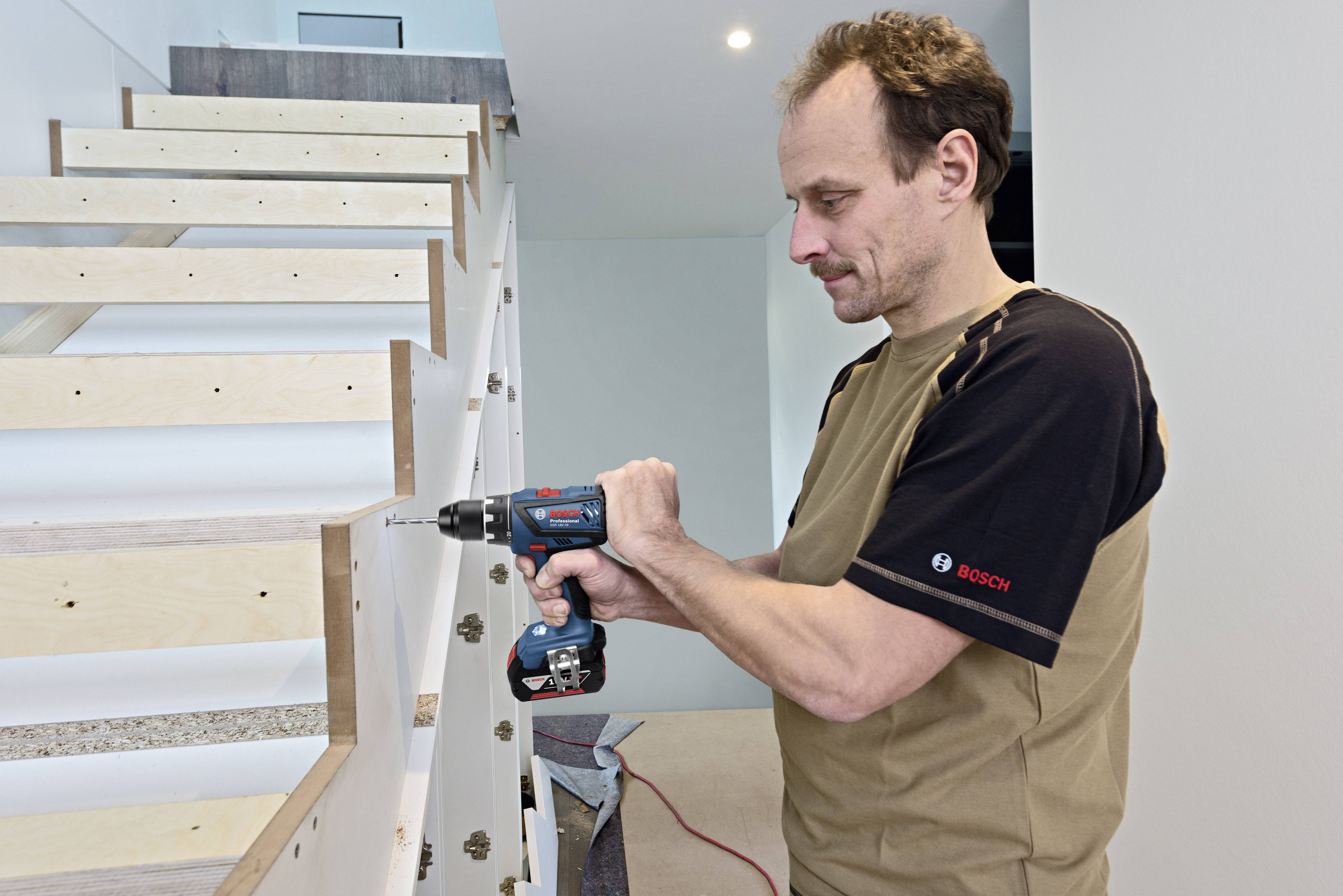 A man is using an electric drill to assemble a wooden staircase. He is wearing a T-shirt and is standing inside a building.