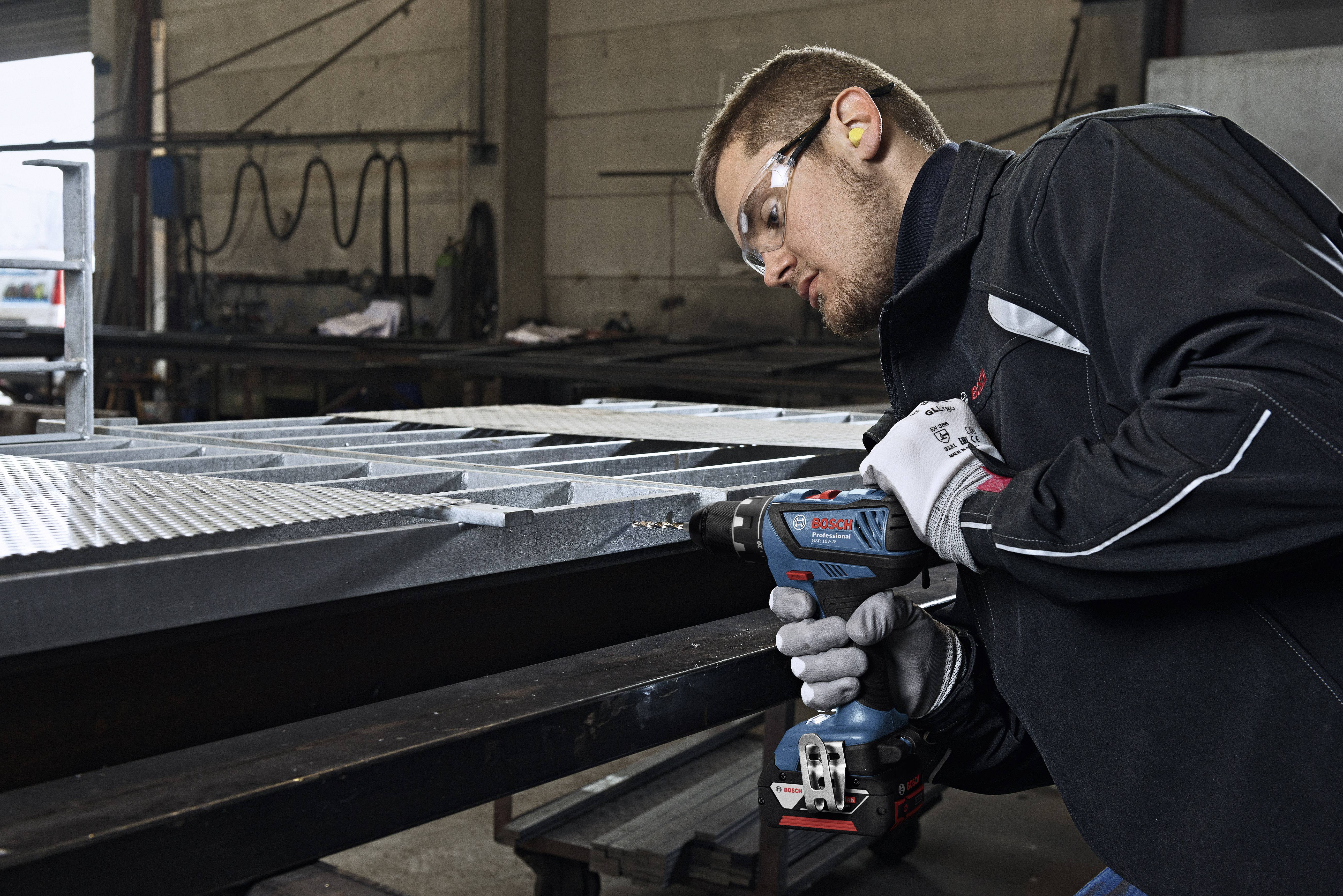 A man in workwear is drilling into metal with an electric drill. He is wearing safety glasses and ear plugs in a workshop.