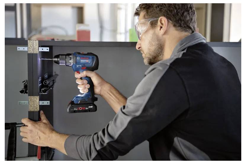 A person wearing safety goggles uses a cordless drill to secure a metal hinge onto a piece of wood, focusing intently on the task.