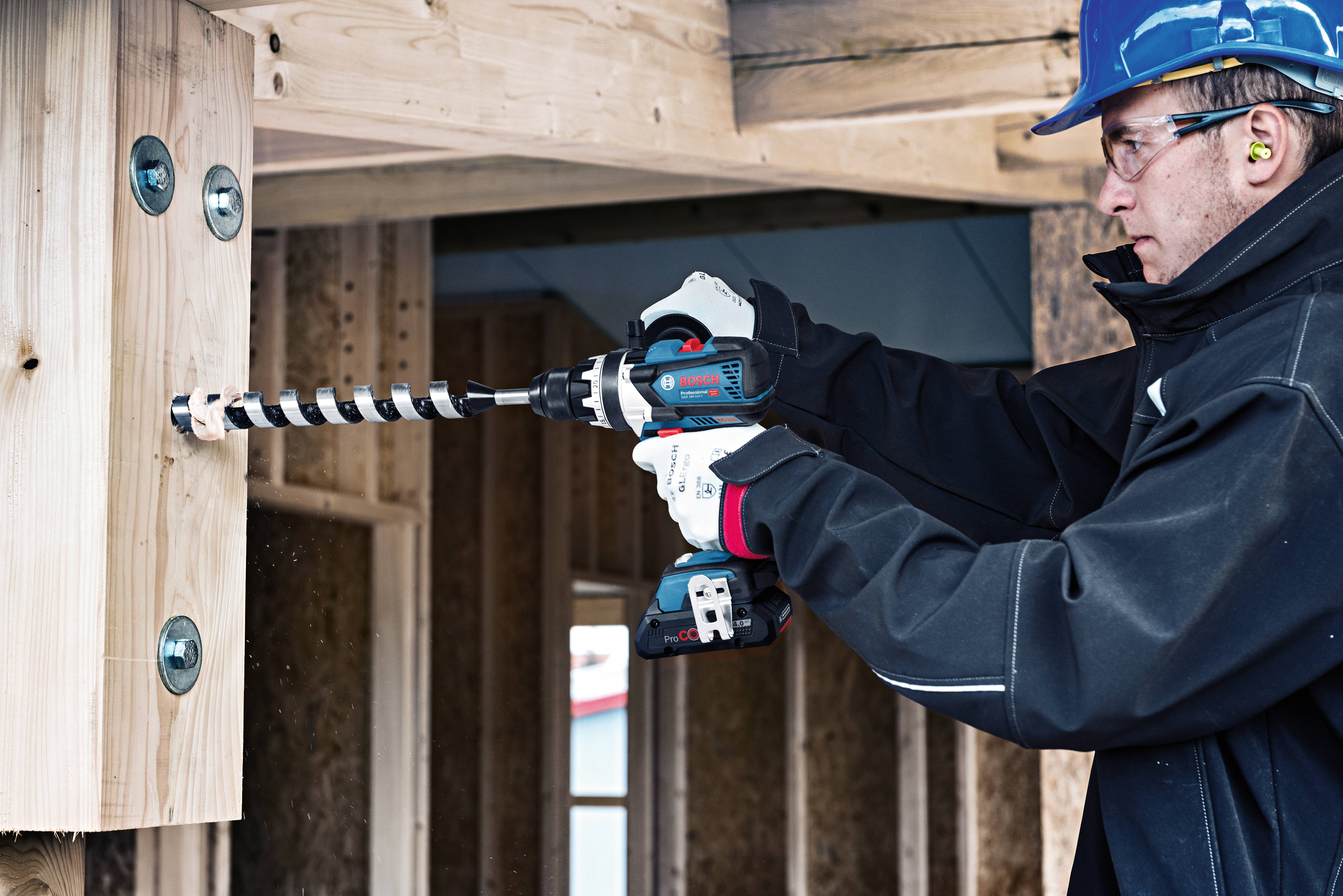 A construction worker is drilling a hole in a wooden beam using a cordless drill. He is wearing protective clothing, a hard hat, and ear protection.