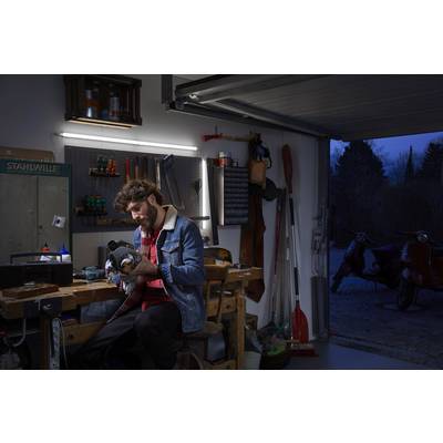 A person is working at a table in a garage, holding a tool. Workshop equipment and mopeds can be seen in the background.