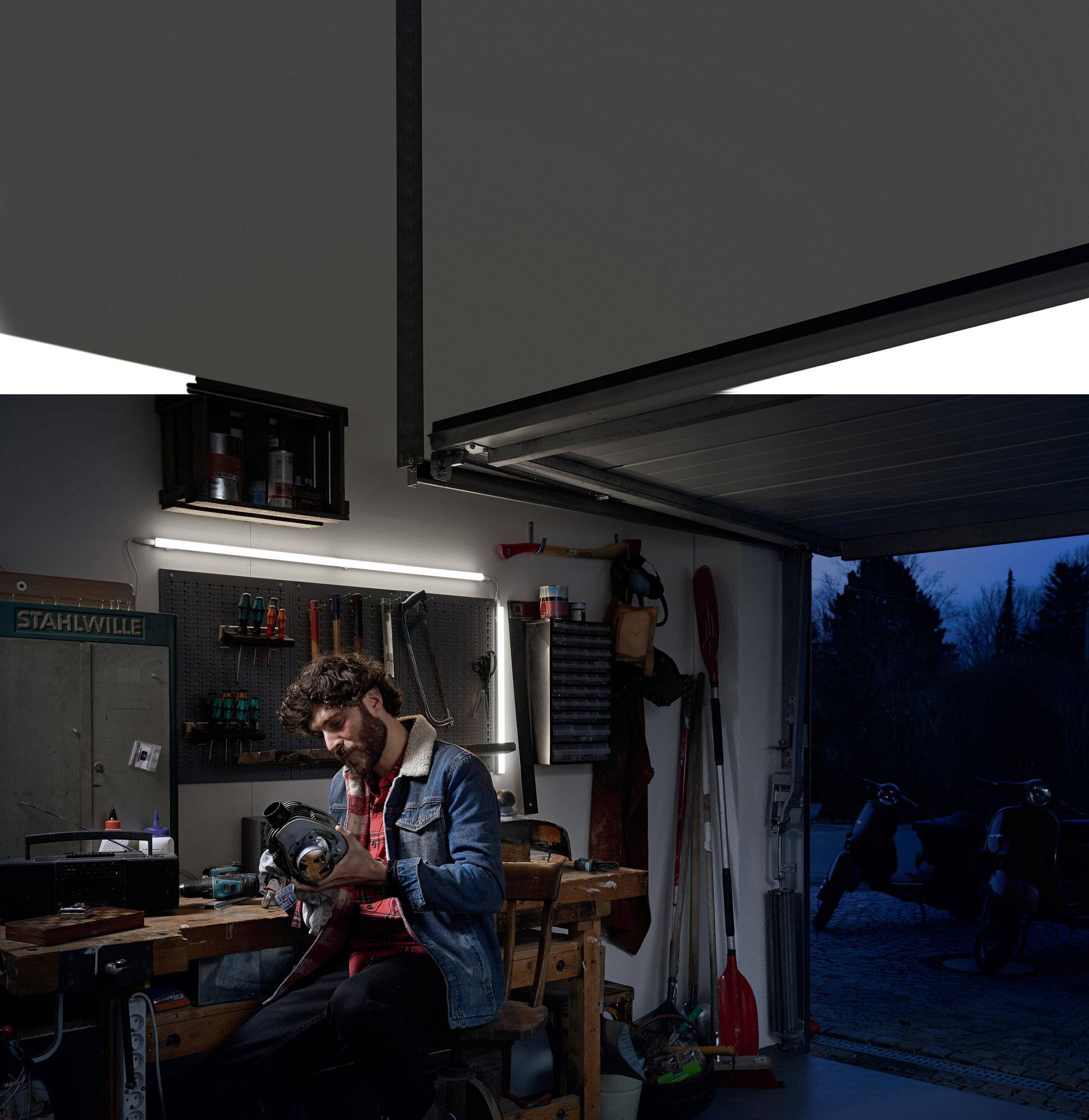 A man is sitting in a garage and working on a tool. The garage is dimly lit, and it is night outside.