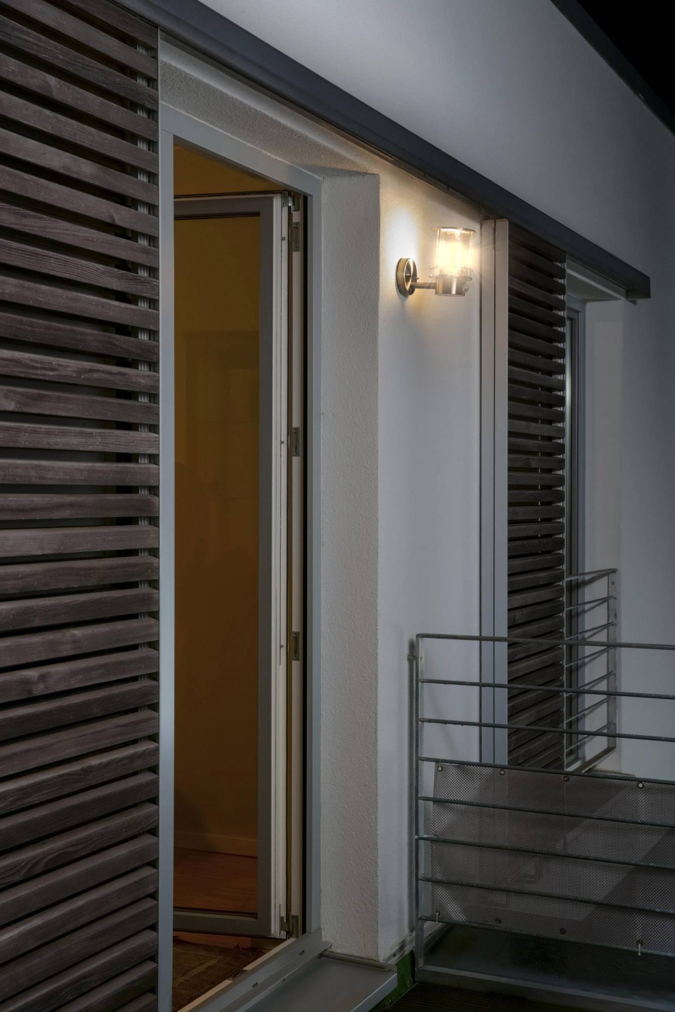Open terrace door at night, illuminated by an outside light; bright, modern house facade with wooden elements.<br><br>Translation to British English:<br>Open patio door at night, lit by an exterior lamp; bright, contemporary house exterior with wooden features.