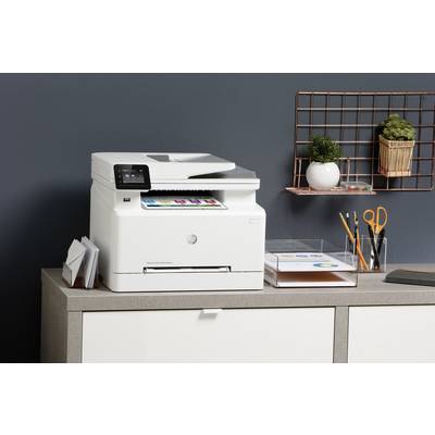 A white printer on a cupboard is printing a coloured piece of paper. Next to it are office supplies such as pens in a holder.