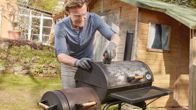 A person is barbecuing outdoors with a large barbecue, using barbecue tongs. A wooden shed is visible in the background.