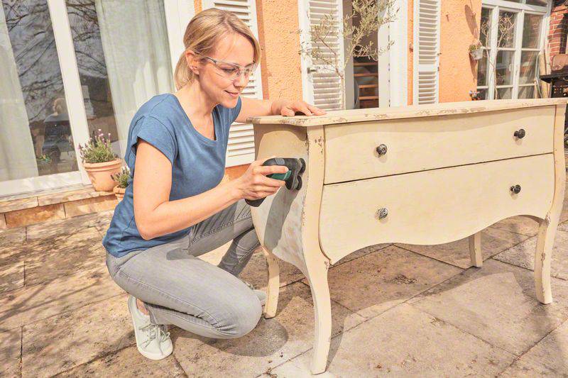 A person is sanding the surface of an antique chest of drawers outdoors using an electronic sanding device.