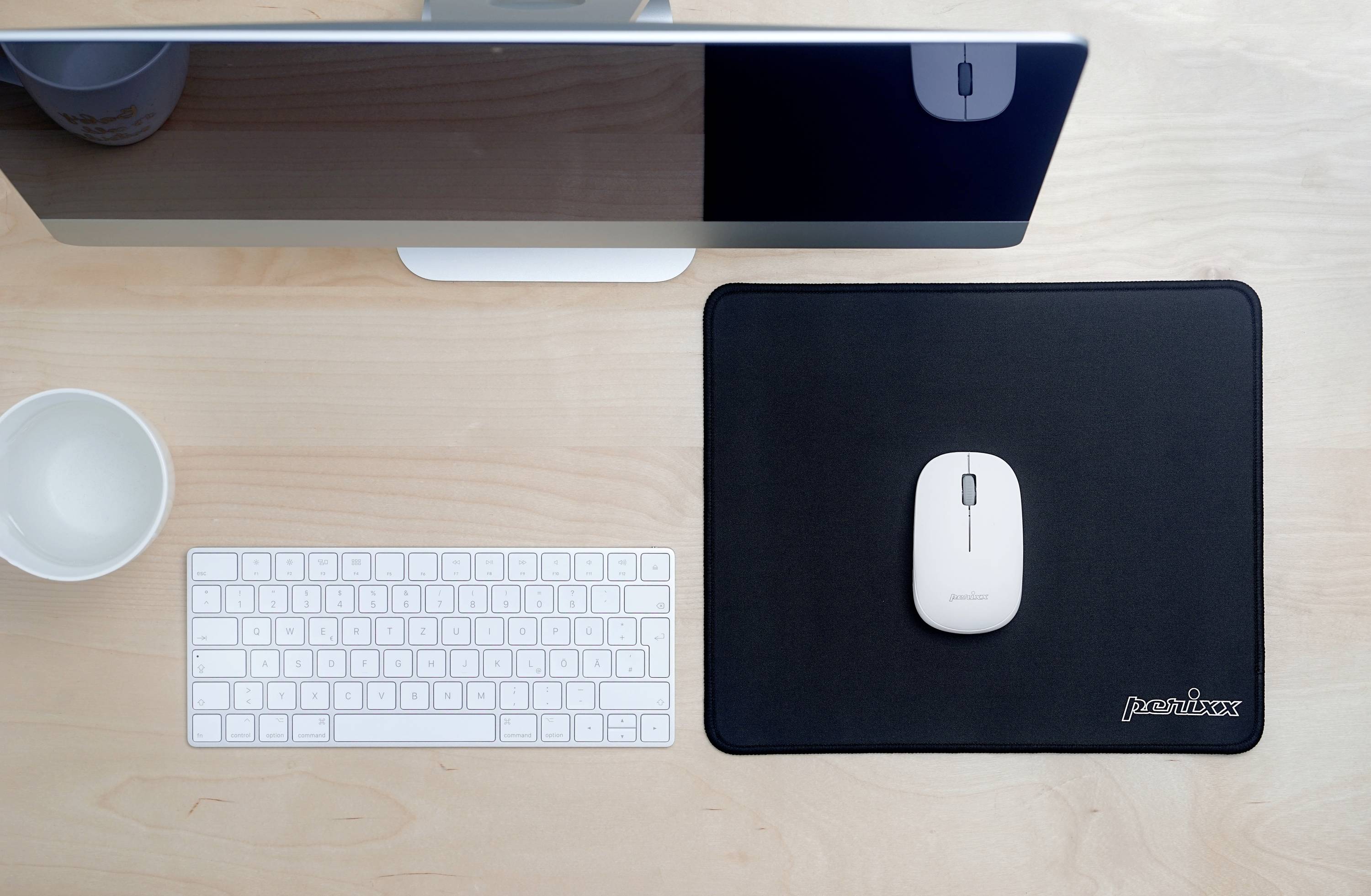 A computer monitor, keyboard and mouse on a clean wooden desk. A cup is positioned to the left of the keyboard.