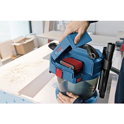 A person is inserting a red battery into a blue, portable vacuum cleaner. Cardboard boxes and furniture can be seen in the background.