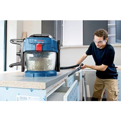 A man is cleaning a kitchen worktop with an industrial vacuum cleaner. The vacuum is blue and sits on the work surface. The man is wearing safety glasses.