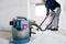A man is cleaning a concrete floor with a Bosch construction vacuum cleaner in a bright room. The vacuum cleaner is blue and transparent.