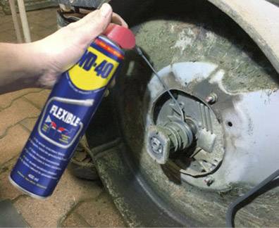A hand sprays oil from a can onto a rusty machine part. The can, labelled 'WD-40 Flexible', has a red spray nozzle and a flexible tube.