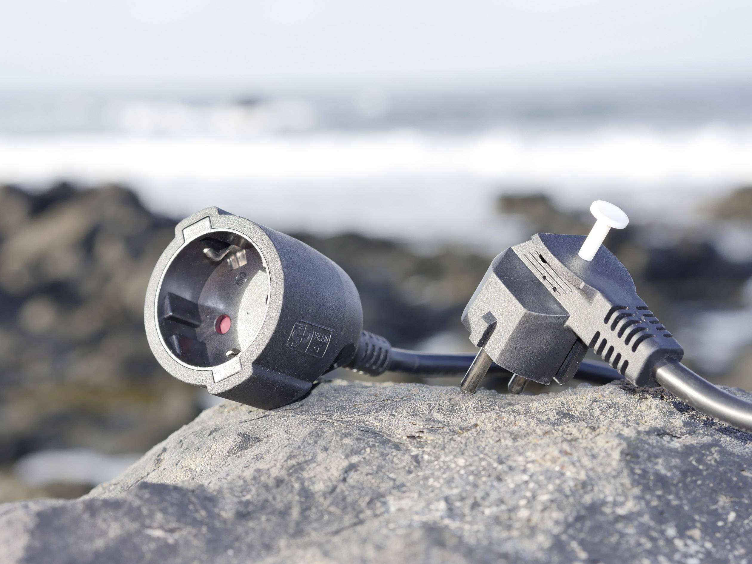 A European plug socket and plug are lying on a rock with a blurred sea background.