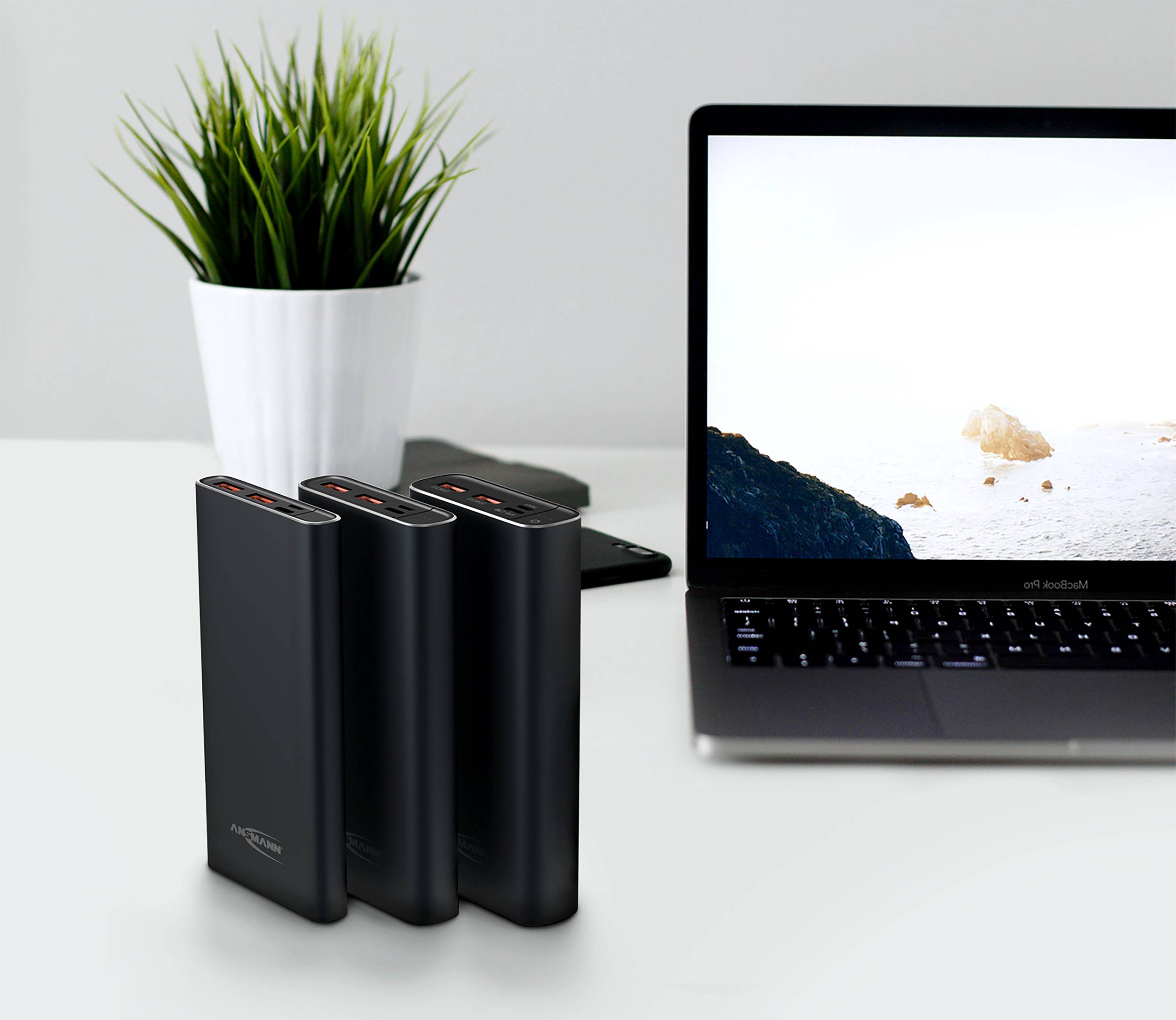 Three black power banks are lined up next to each other on a desk beside an open laptop with a landscape screensaver.