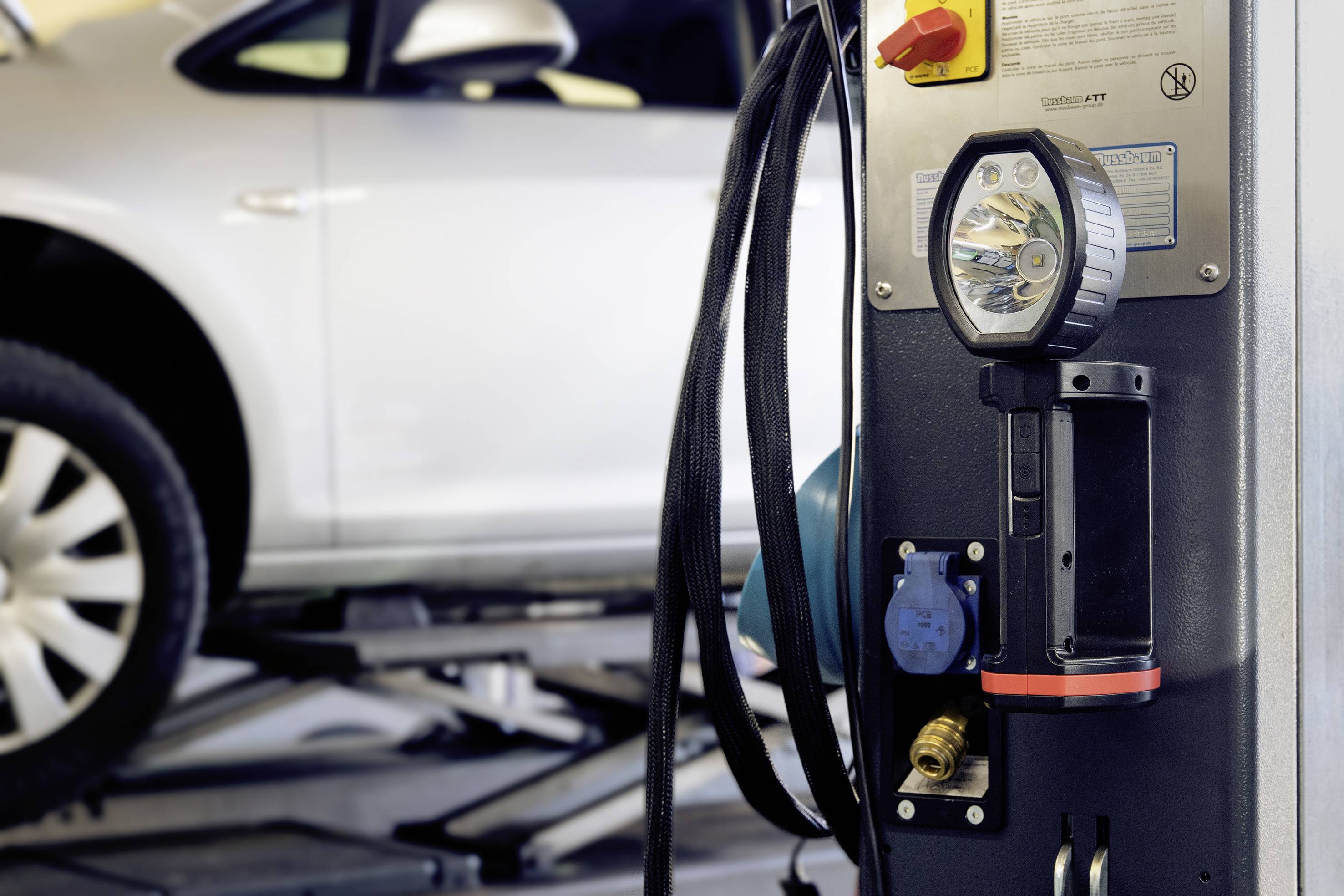 A car is positioned on a car lift in a garage workshop. In the foreground, a tyre inflation station with hoses and gauges is visible.