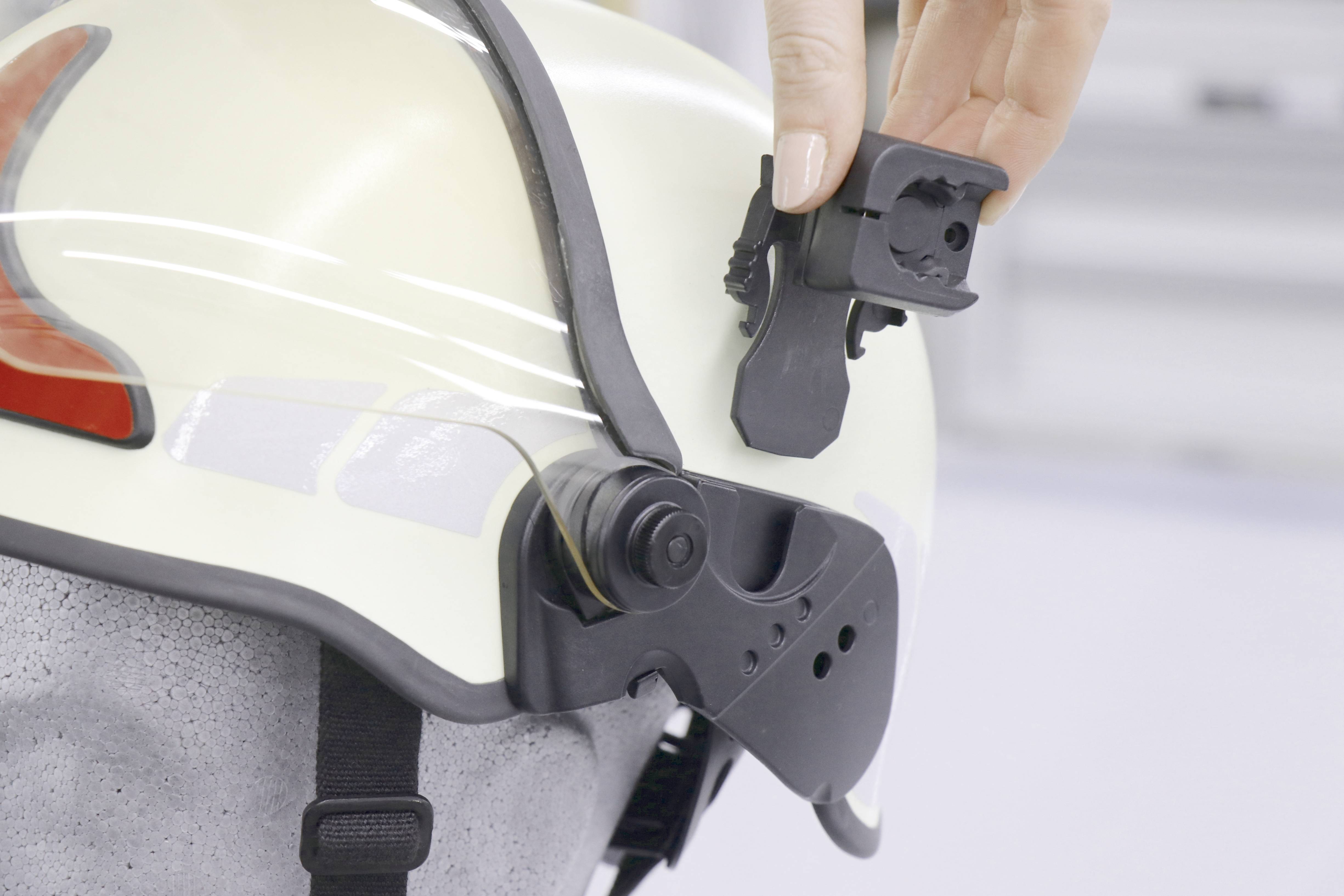 A hand points to a black helmet mount on a white safety helmet. The accessory appears removable and designed for adjusting the helmet.