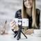 A smiling person is taking a selfie with a smartphone mounted on a tripod. Blurred office supplies are visible in the background.