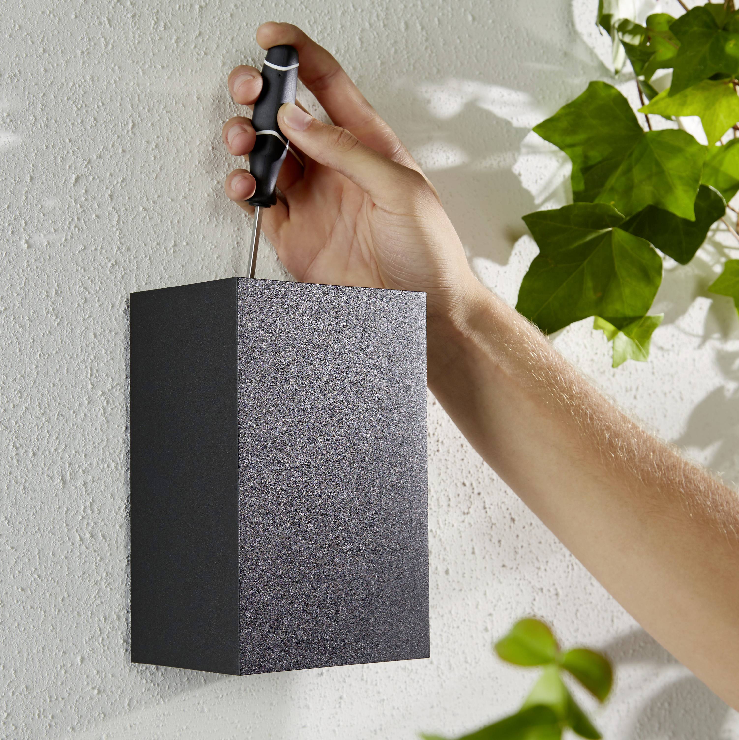 A hand is mounting a black lamp on a white wall with a screwdriver; green plants in the background.
