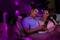 A couple sit close together on a sofa, looking cheerful and sharing a mobile phone screen. The lighting is purple.